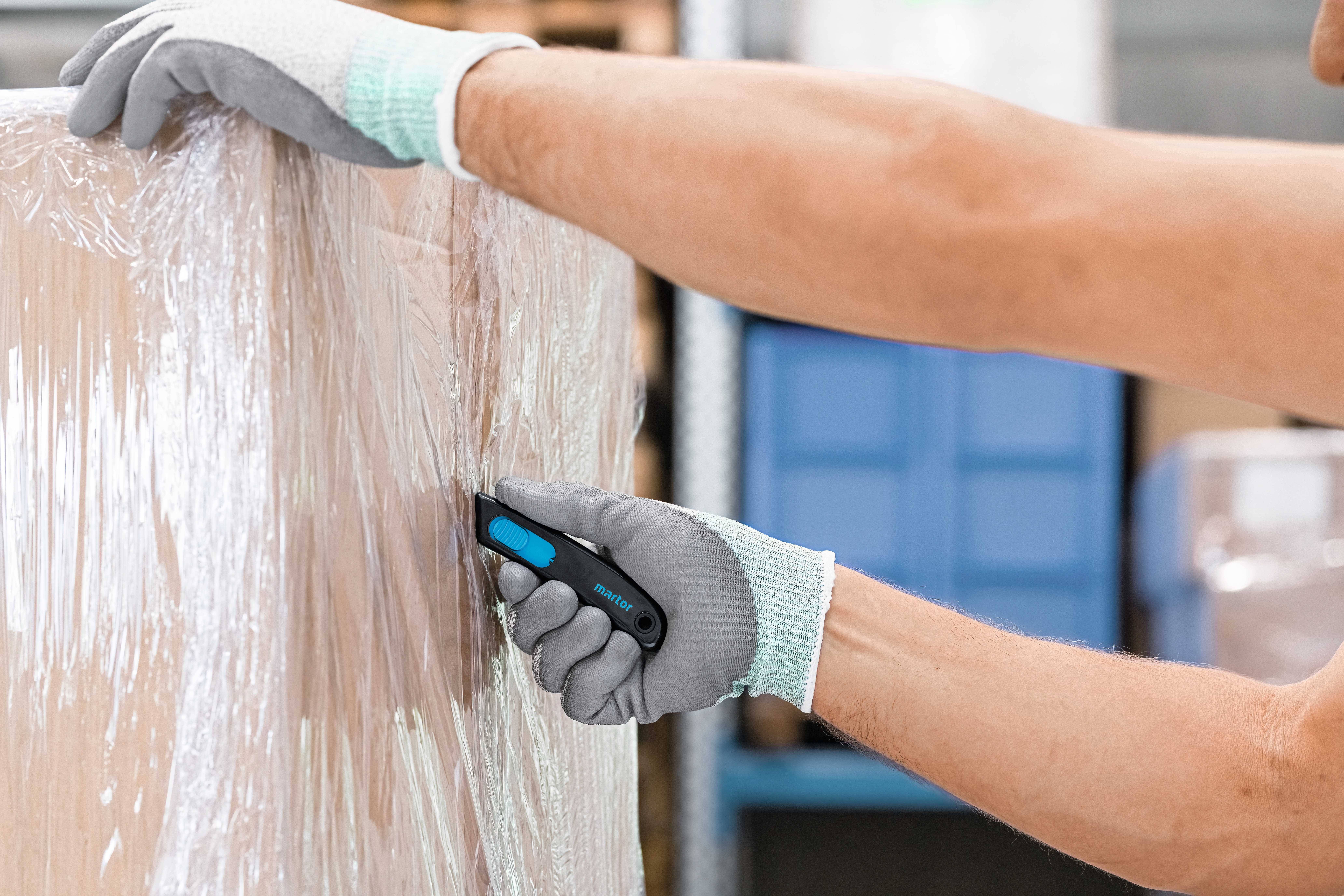 Eine Person trägt Handschuhe und schneidet mit einem Sicherheitsmesser die Plastikfolie von einer Palette ab.