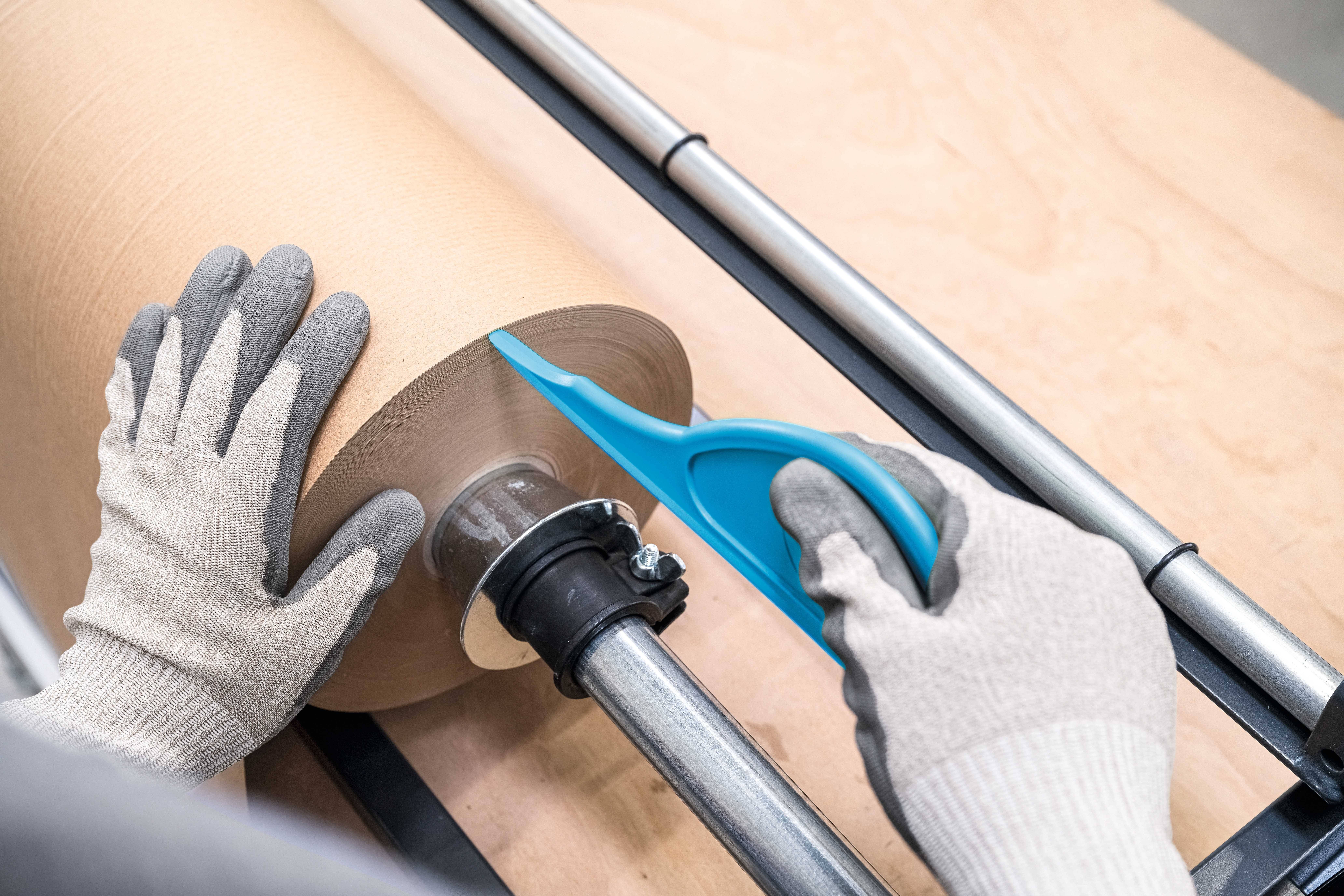 Eine Person schneidet mit einem blauen Messer einen großen Papierrollenkern ab. Sie trägt graue Handschuhe für Schutz.