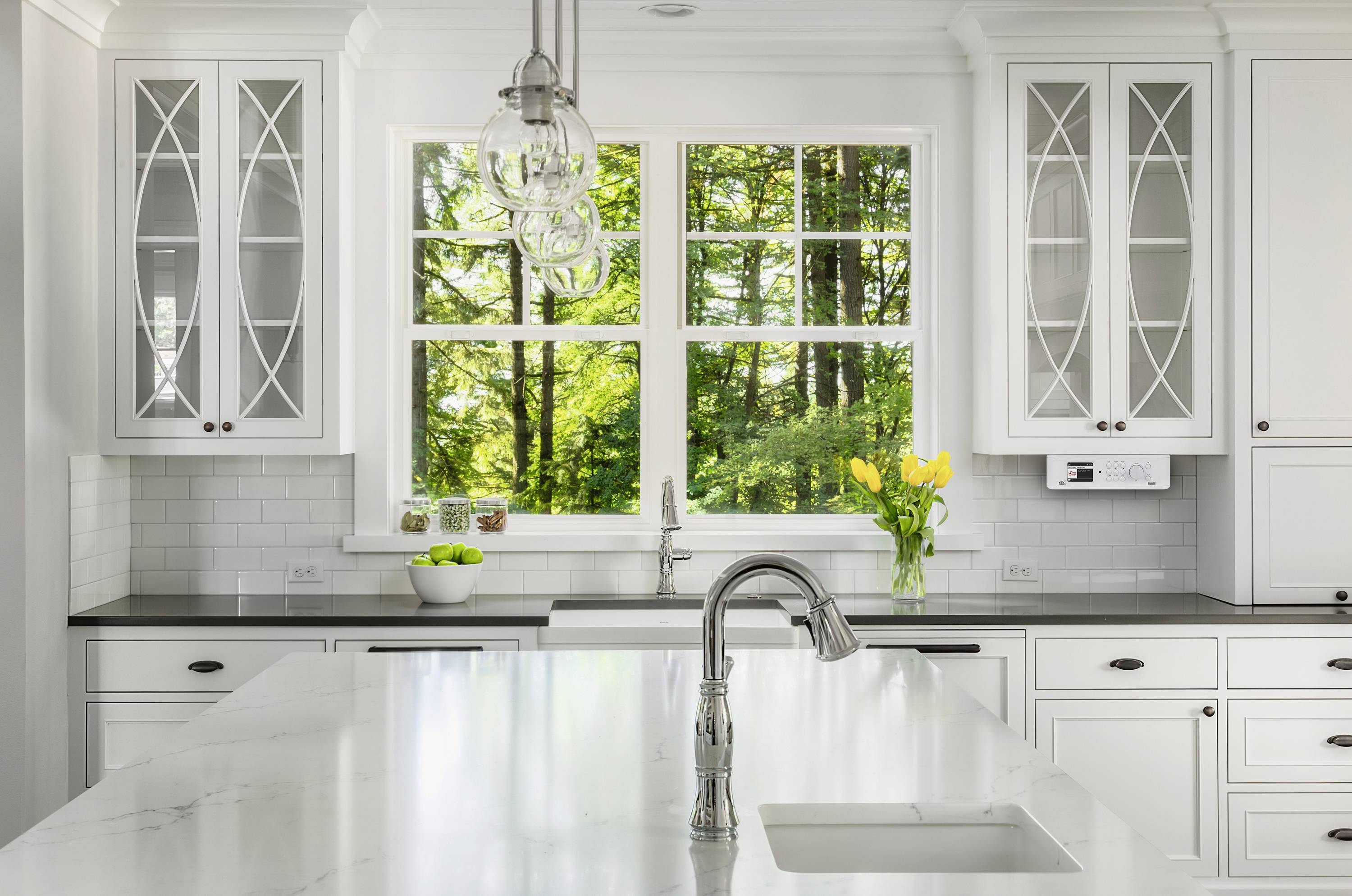 Elegante, moderne Küche mit weißer Ausstattung und großer Fensterfront. Draußen grüner Wald sichtbar. Vase mit gelben Tulpen auf der Theke.