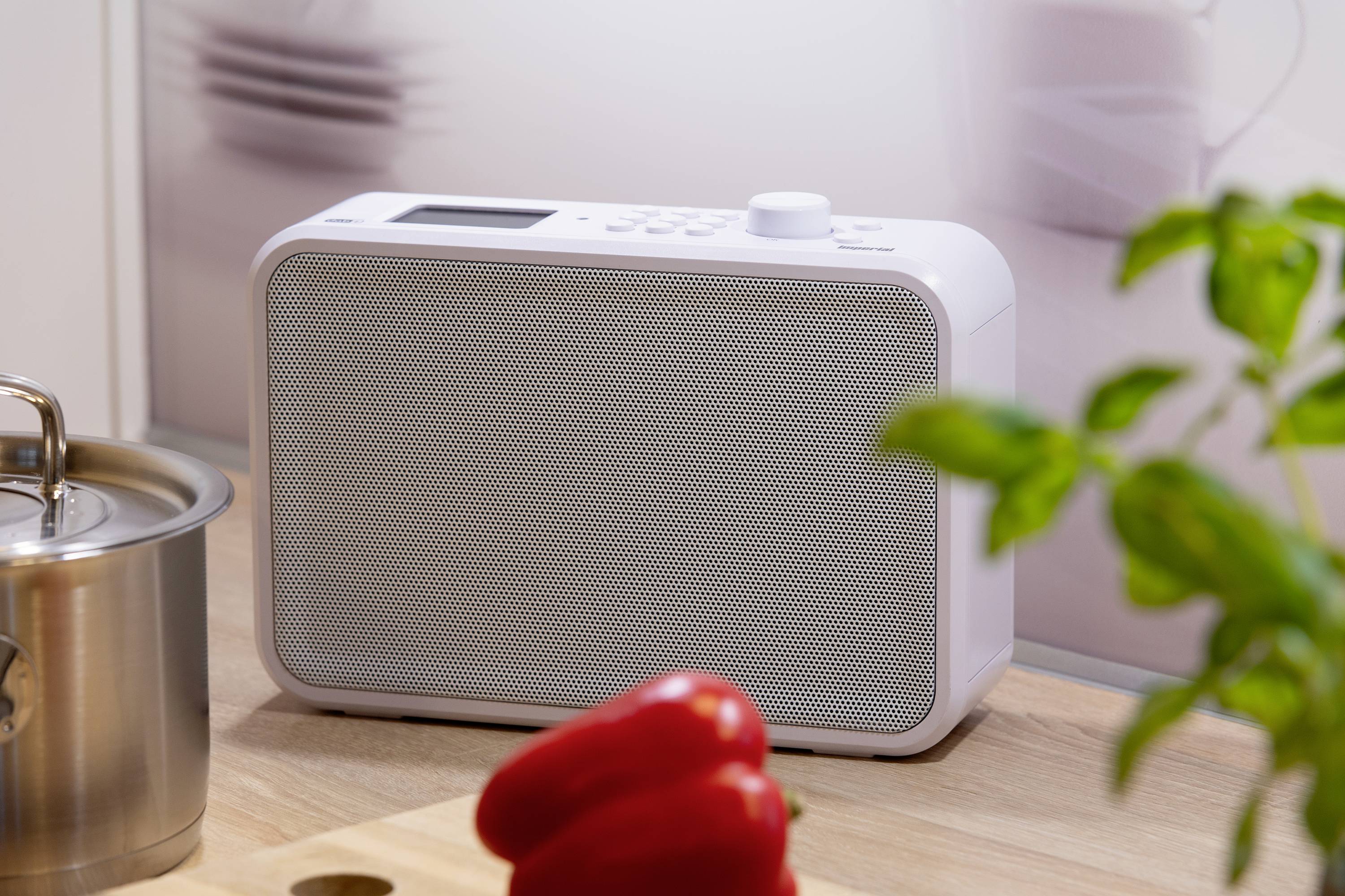 Ein weißes Radio steht auf einer Küchenarbeitsplatte neben einem roten Paprika und einer Topfpflanze, im Hintergrund unscharf eine Pfanne.