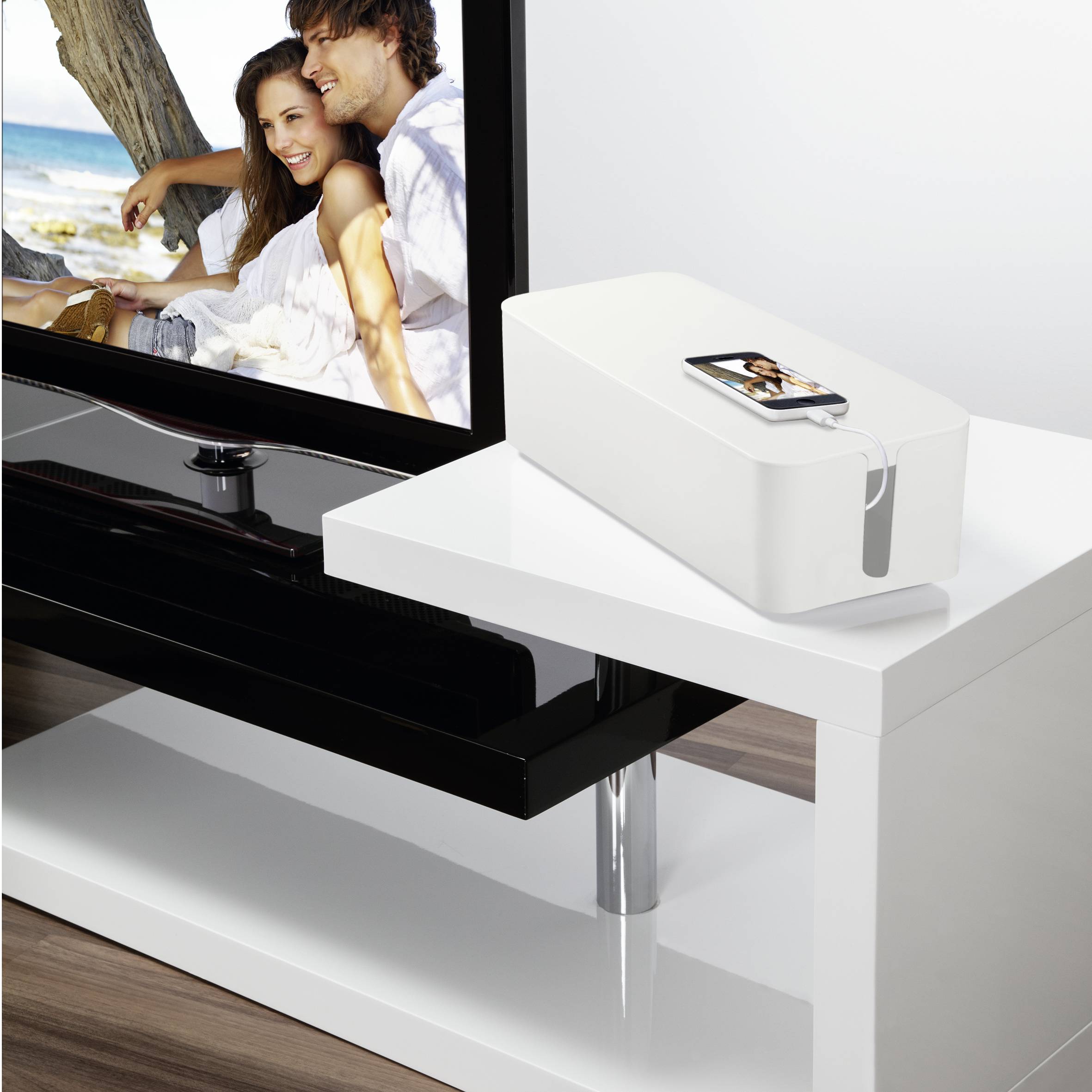 Ein weißer Tisch mit einer Kabelbox darauf, neben einem Flachbildfernseher, der ein Paar am Strand zeigt.