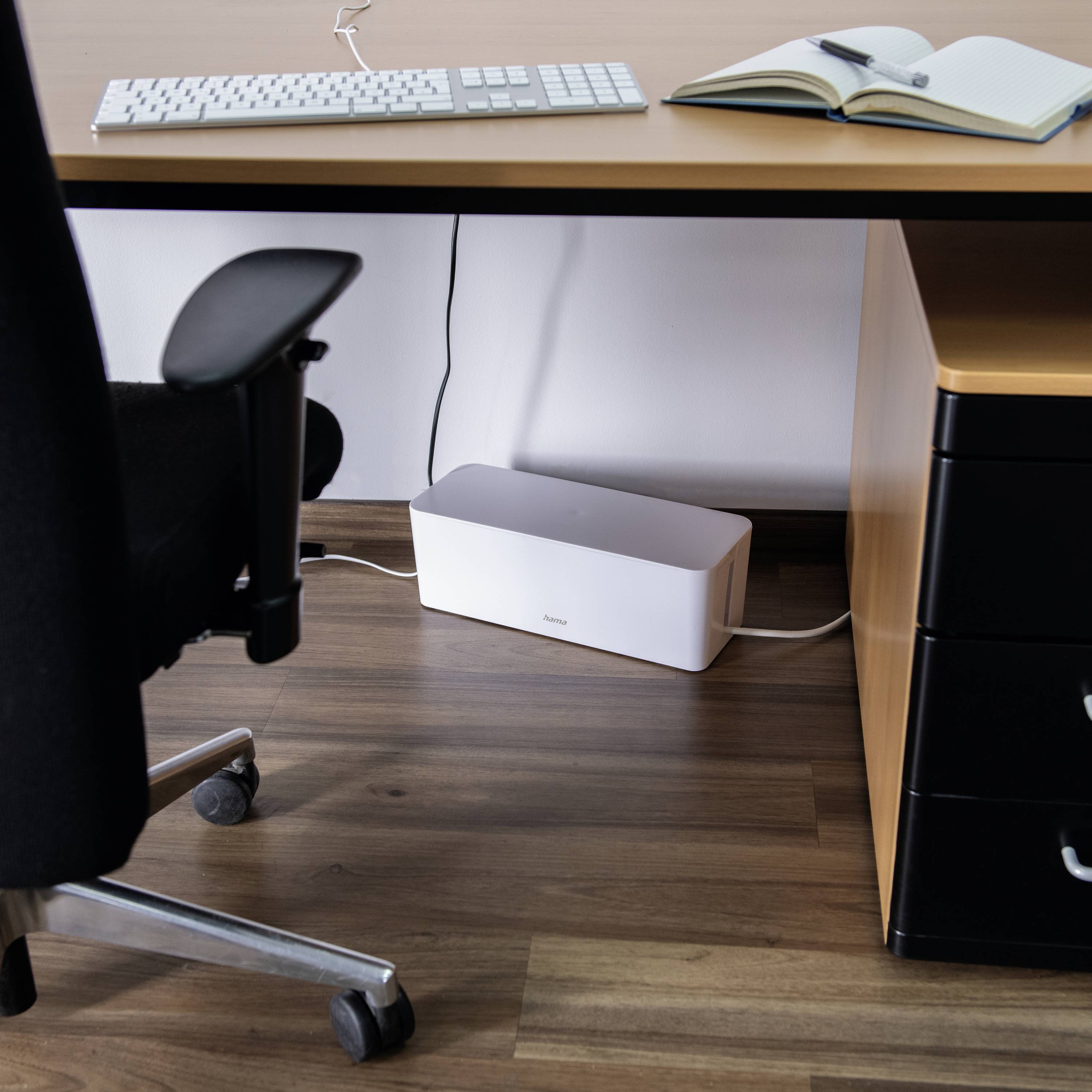 Ein Schreibtisch mit einem Computer, Tastatur, Notizbuch und Stift. Darunter steht ein Router neben einer Schubladeneinheit auf Holzboden.