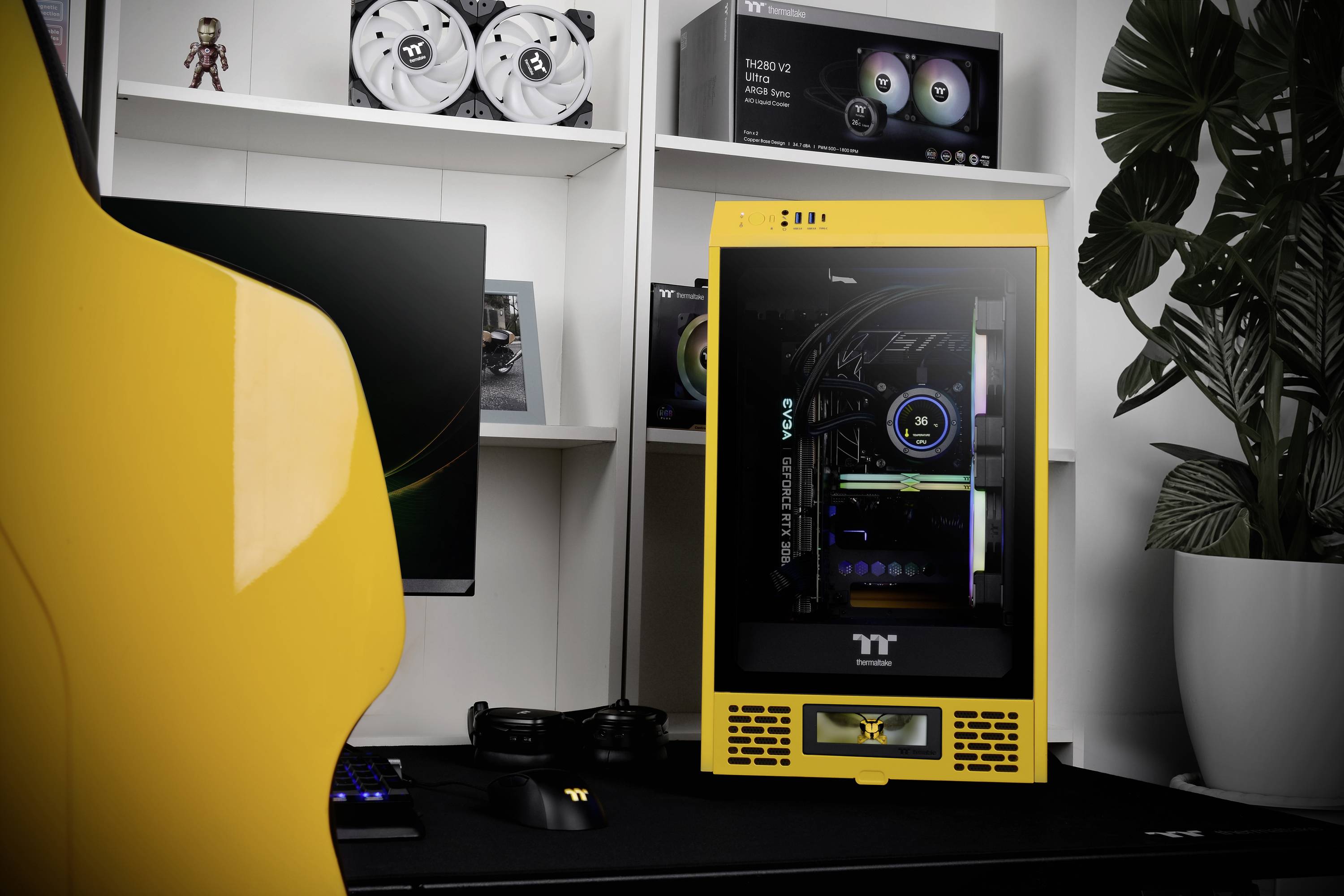 Ein gelber Gaming-PC mit durchsichtigem Gehäuse zeigt innenliegende Hardware. Im Hintergrund ist ein weißes Regal mit Computerteilen und grüner Pflanze zu sehen.