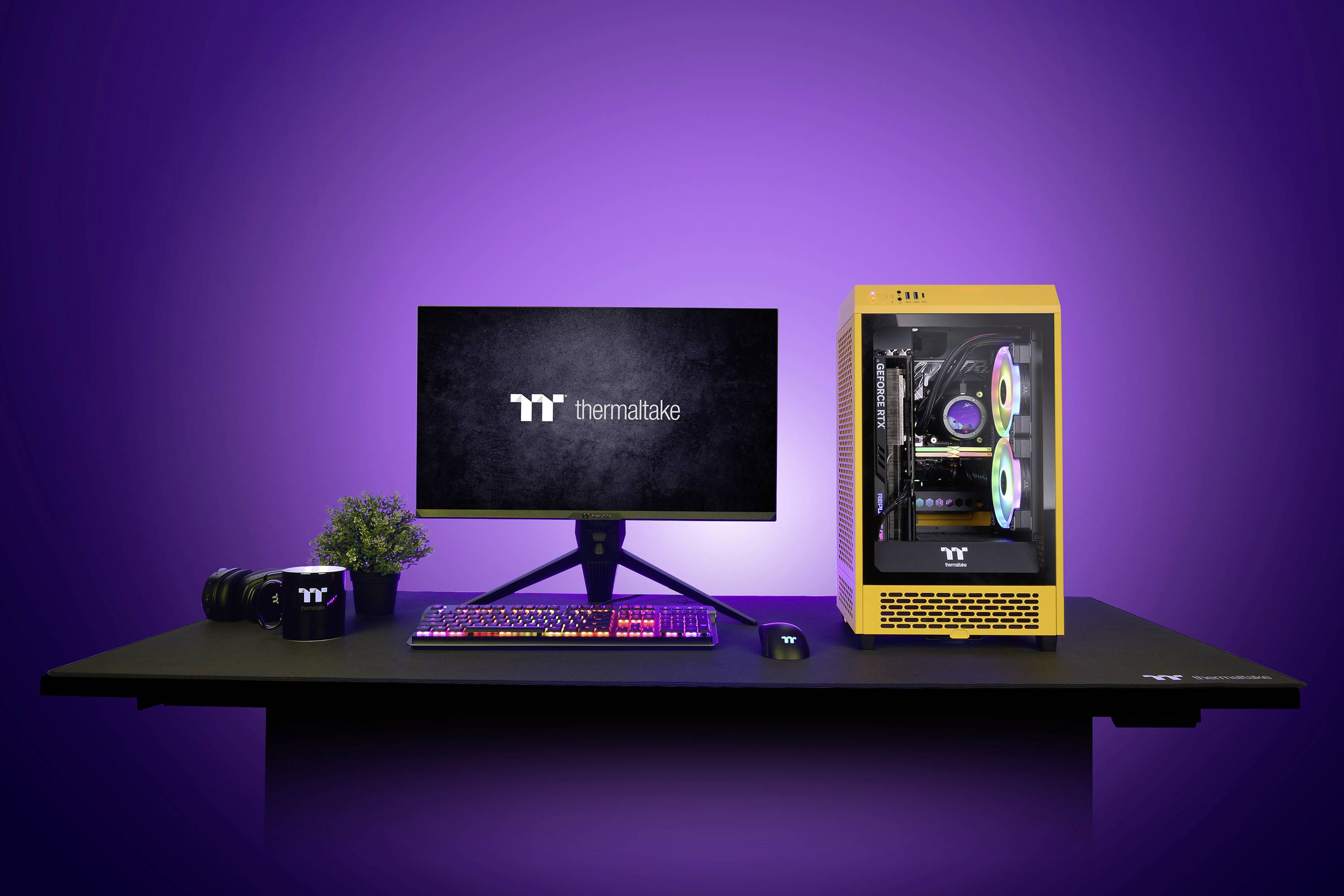 Ein Schreibtisch mit einem Computer-Setup. Monitor zeigt 'Thermaltake'-Logo, dazu Tastatur, Maus, Kamera, Pflanze und gelbes PC-Gehäuse.