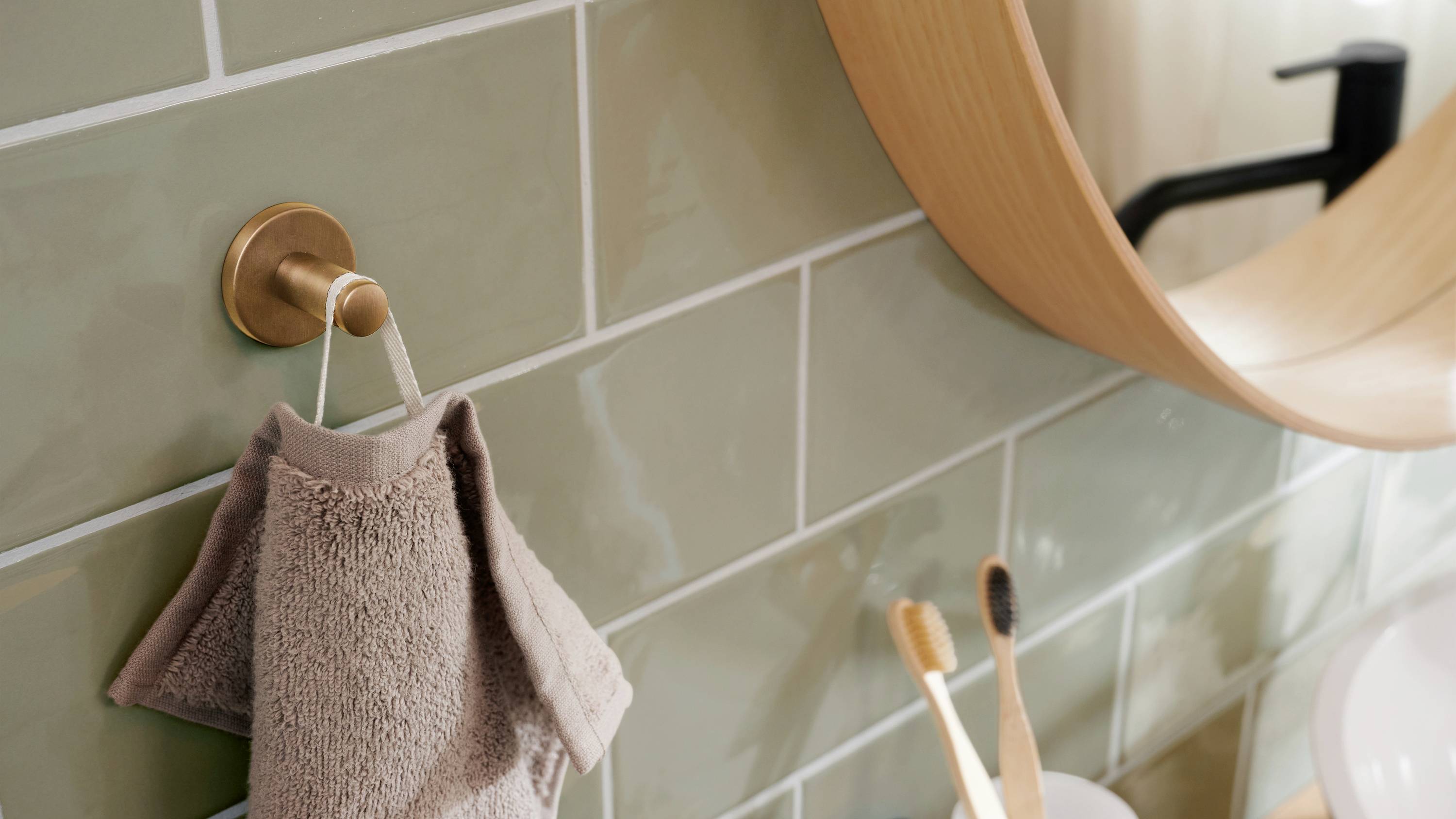 Handtuch hängt an Haken über beigen Fliesen, daneben stehen Zahnbürsten im Becher vor einem runden Holzzimmerspiegel.