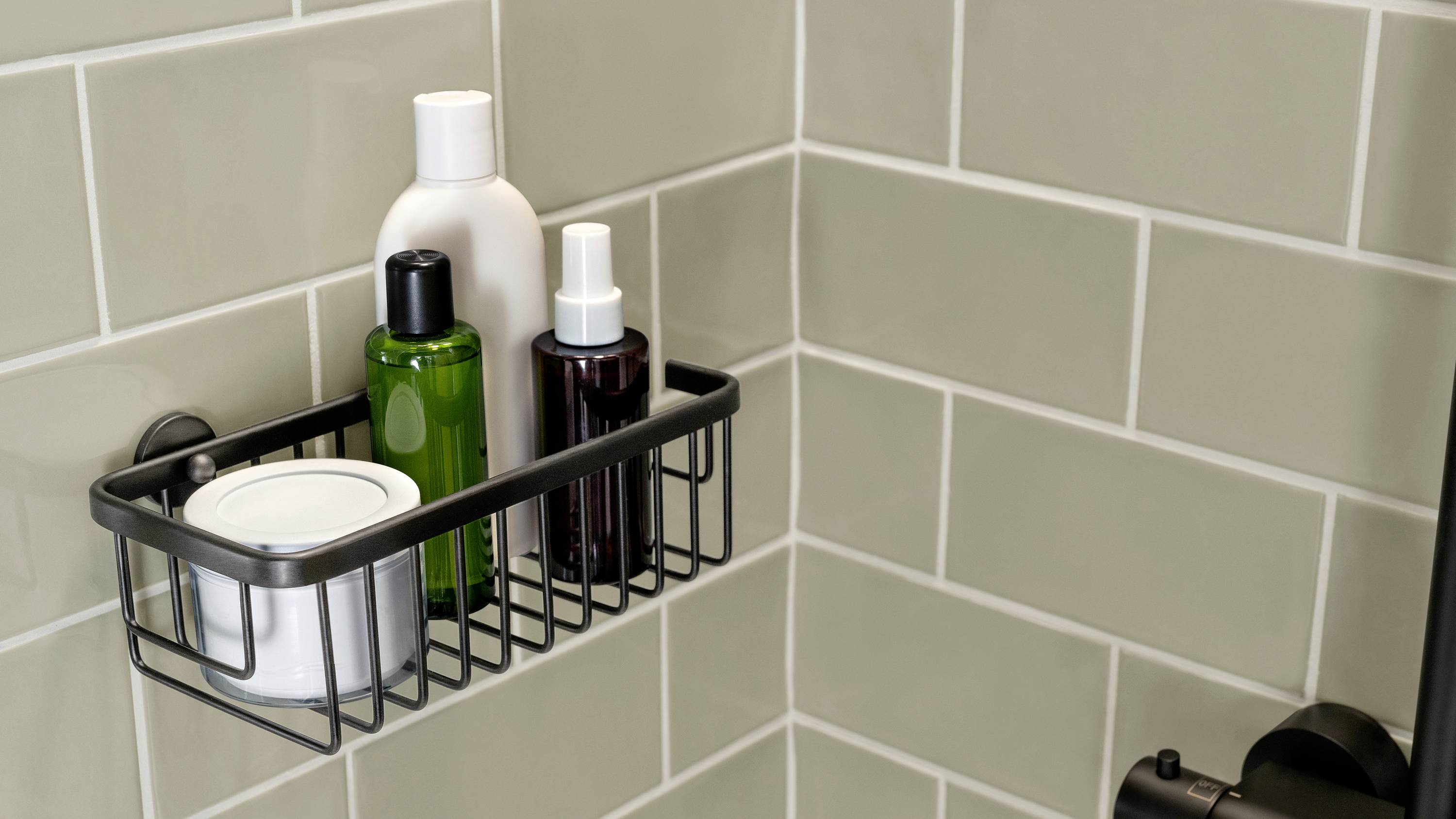 Mehrere Flaschen mit Pflegeprodukten in einem Metallkorb an einer gefliesten Badezimmerwand. Produkte sind weiß, grün und dunkelrot.