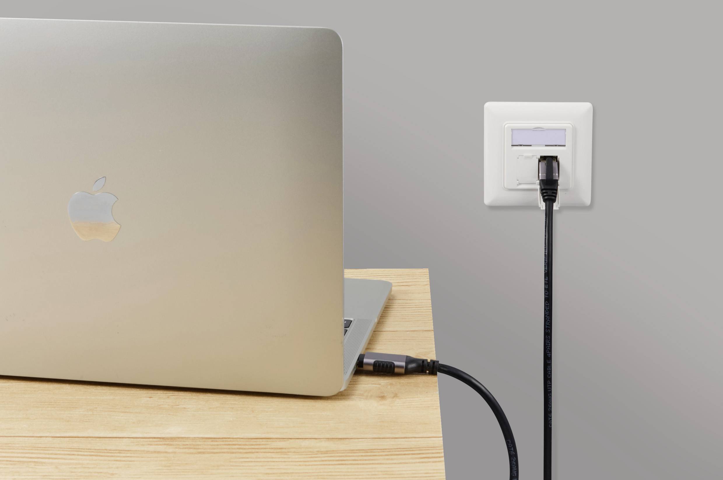 Ein silberner Laptop auf einem Holztisch ist mit einem schwarzen Kabel an eine Steckdose angeschlossen.