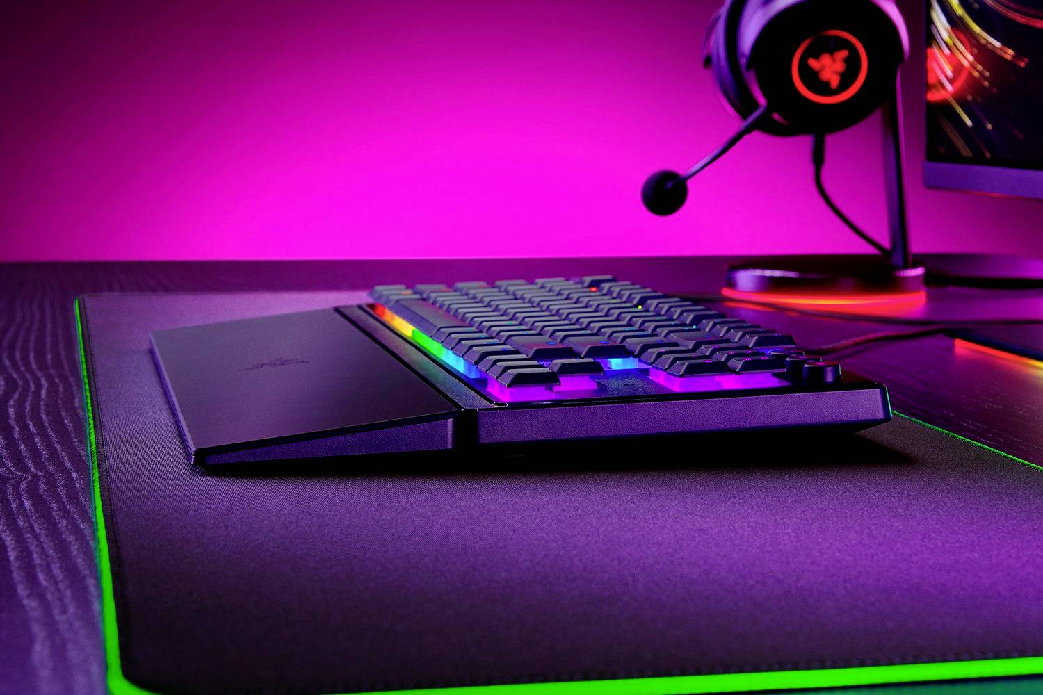 Tastatur mit RGB-Beleuchtung auf Schreibtisch, daneben leuchtendes Gaming-Headset und Monitor mit Hintergrundbeleuchtung in Violett.