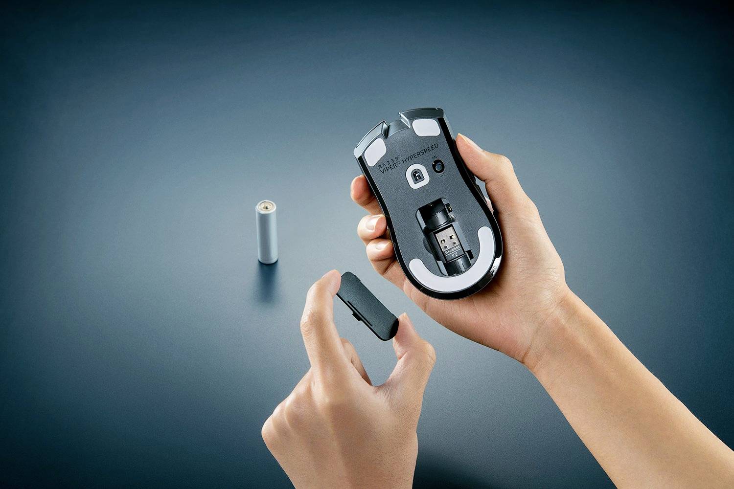 Eine Person hält eine Computermaus ohne Batteriefachabdeckung, während eine Batterie daneben auf dem Tisch liegt.