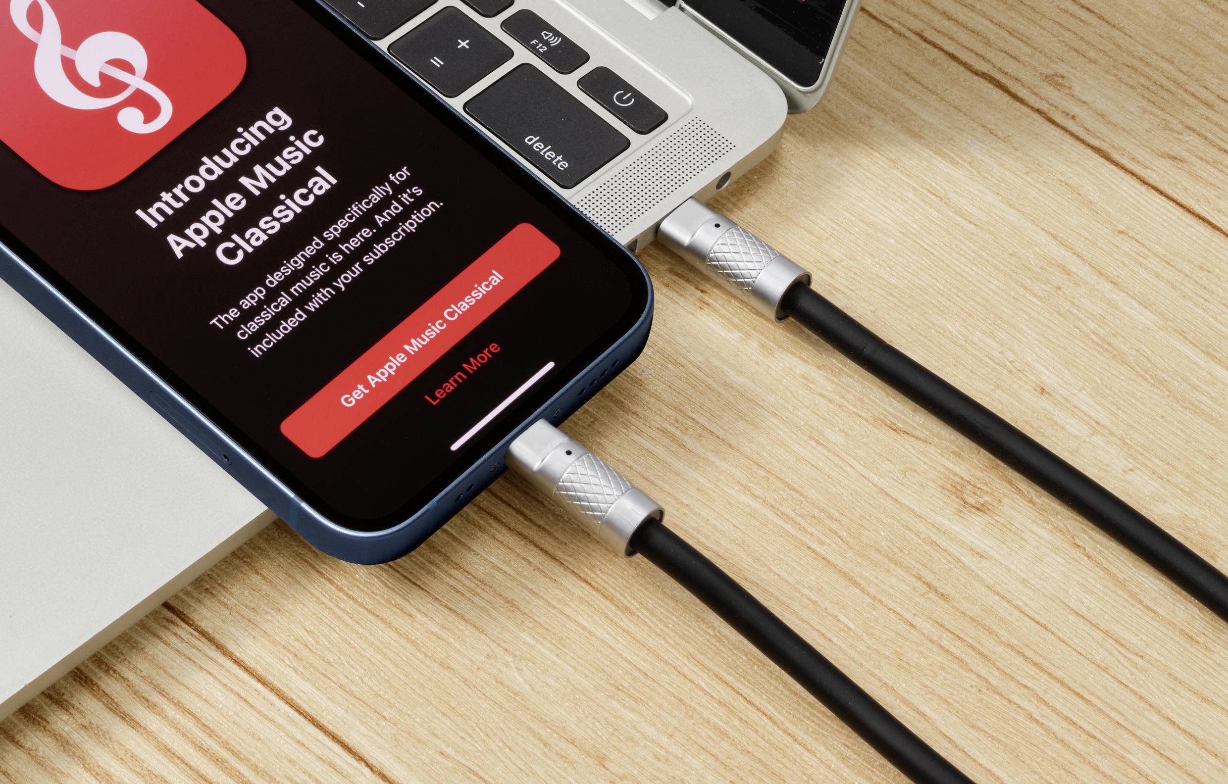 Ein Smartphone auf einem Laptop mit der Anzeige 'Introducing Apple Music Classical', verbunden durch zwei USB-Kabel auf einem Holztisch.