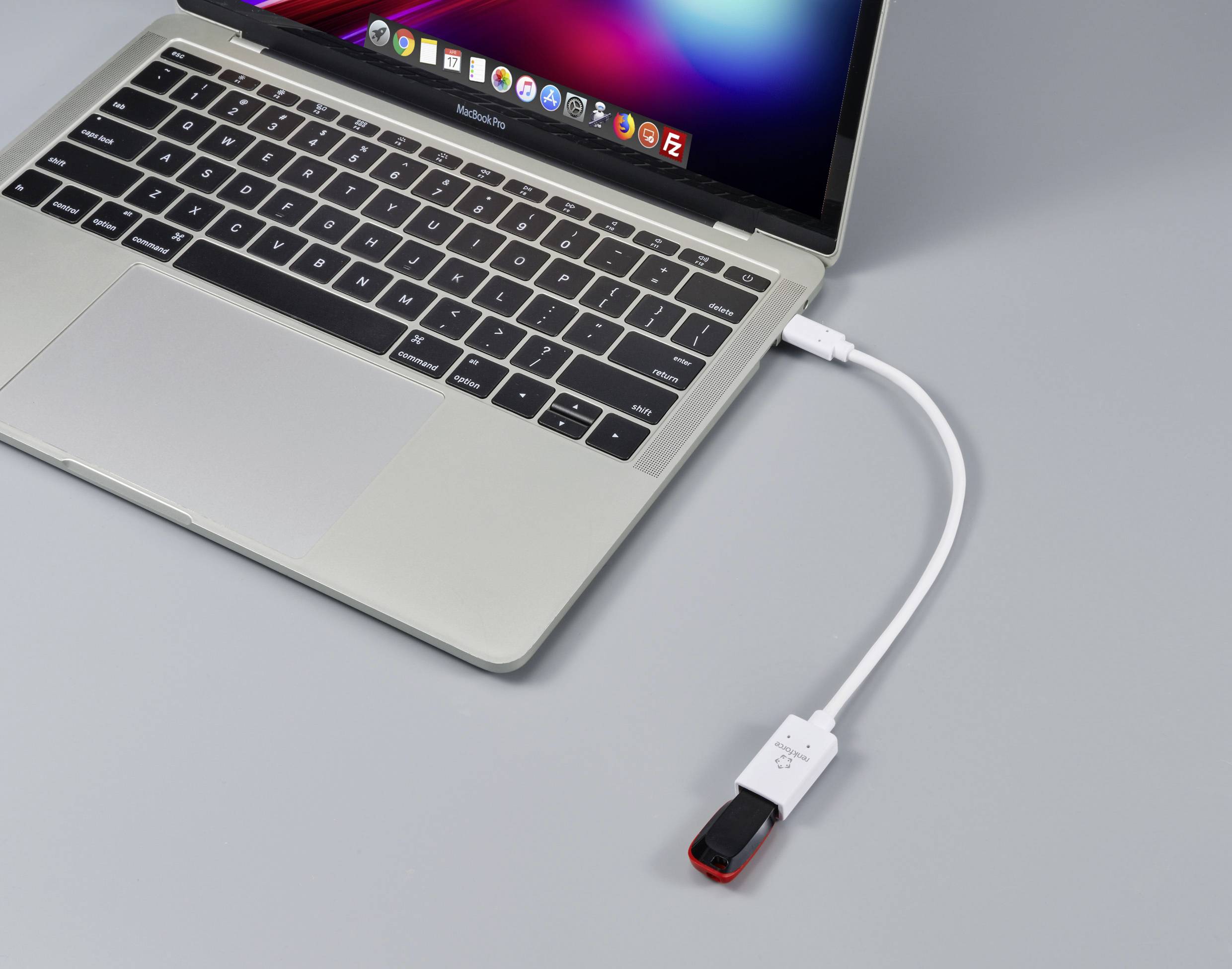 Ein Laptop mit einem offenen Bildschirm ist über ein kurzes weißes Kabel mit einem USB-Stick verbunden, der auf einem Tisch liegt.