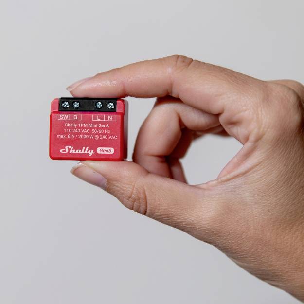 Eine Hand hält ein kleines rotes Gerät namens 'Shelly Gen3' mit elektrischen Anschlussklemmen und technischen Spezifikationen auf der Vorderseite.