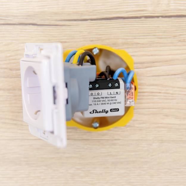Ein intelligenter Wandschalter, teilweise eingebaut, zeigt die Rückseite mit elektrischen Anschlüssen und farbigen Kabeln.