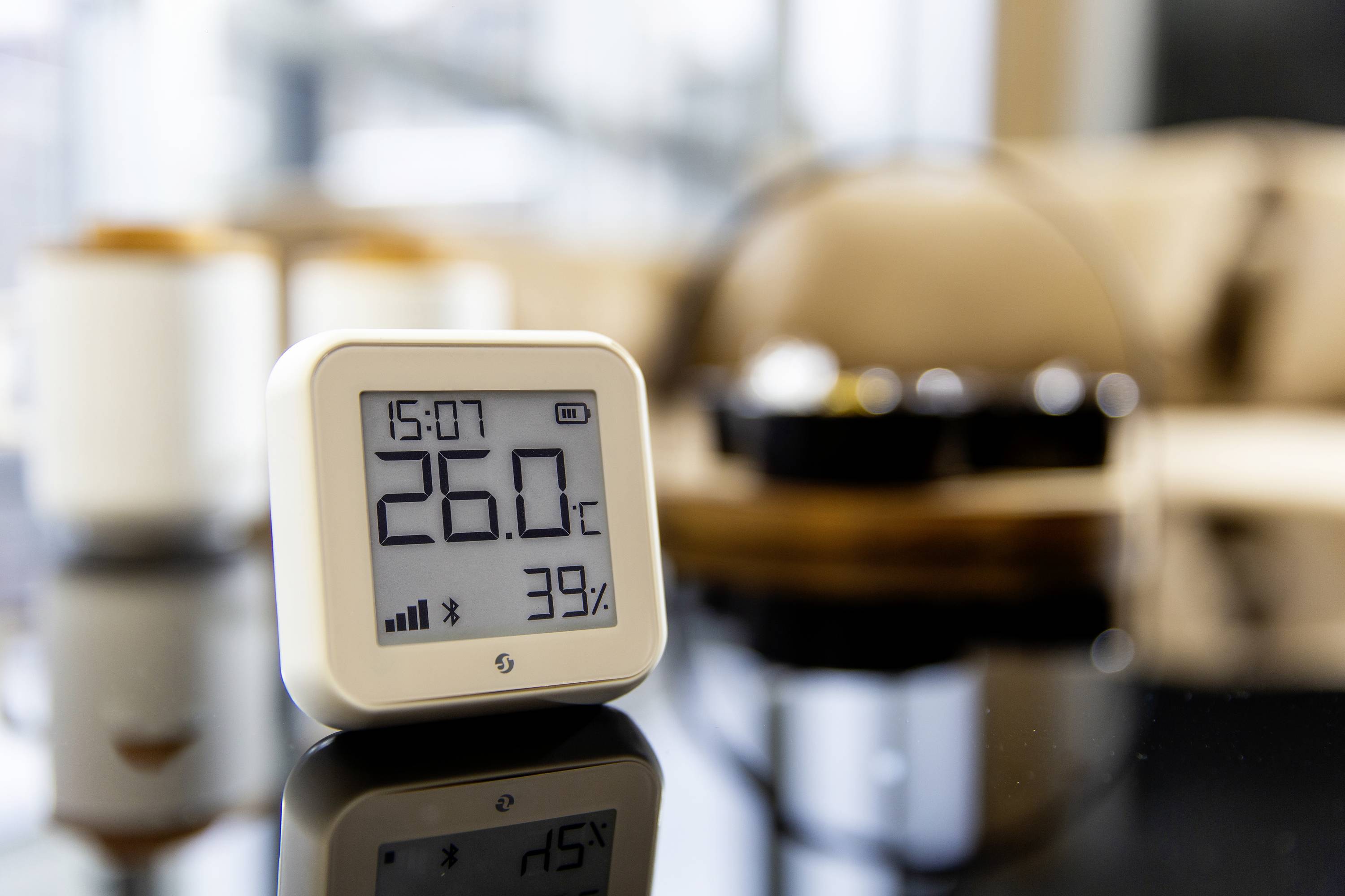 Ein digitales Thermometer auf einem Tisch zeigt 26,0°C und 39% Luftfeuchtigkeit. Im Hintergrund sind verschwommene Wohnmöbel zu sehen.