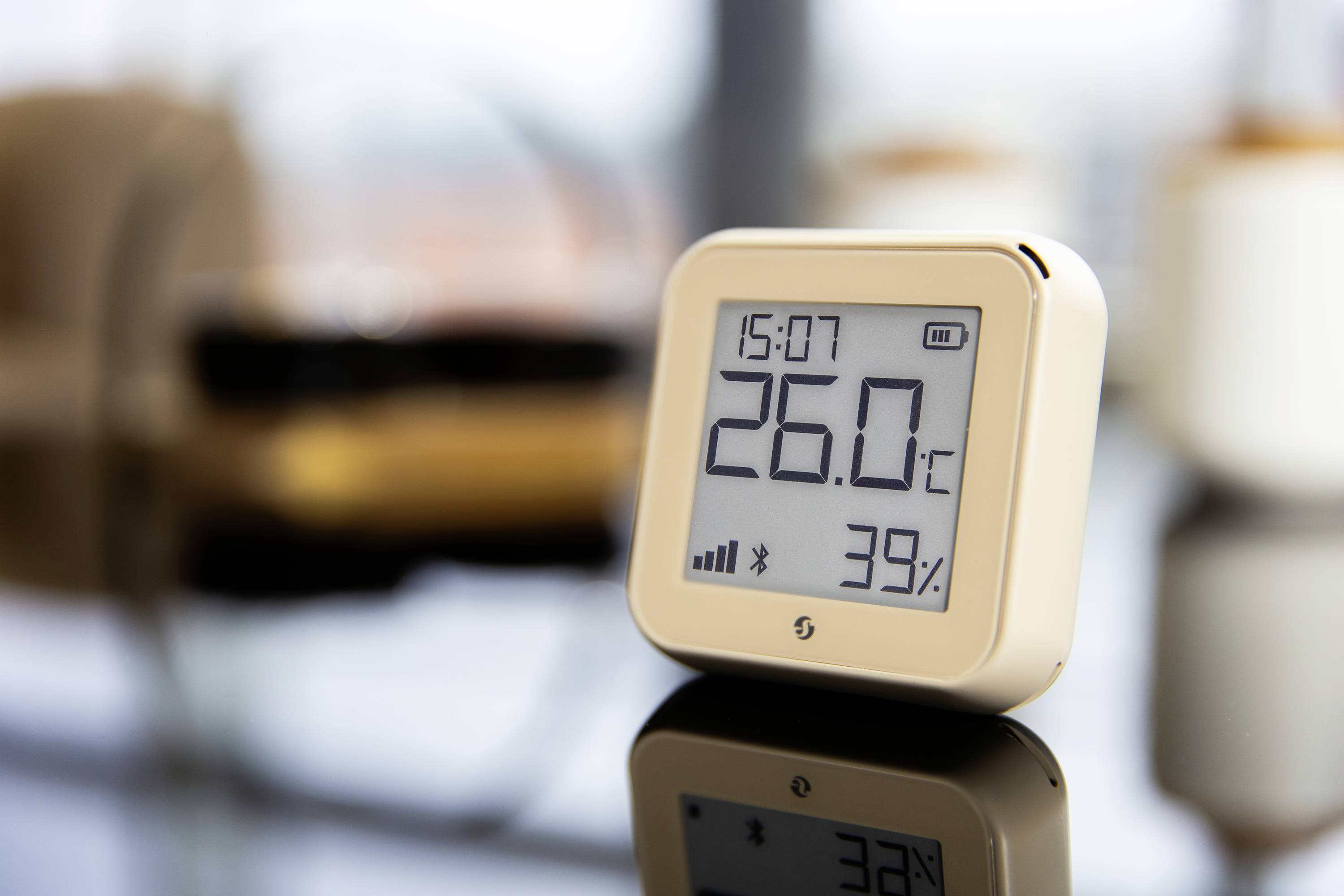 Ein digitales Thermometer zeigt 26,0°C und 39% Luftfeuchtigkeit an. Es steht auf einer spiegelnden Oberfläche in einem modernen Raum.