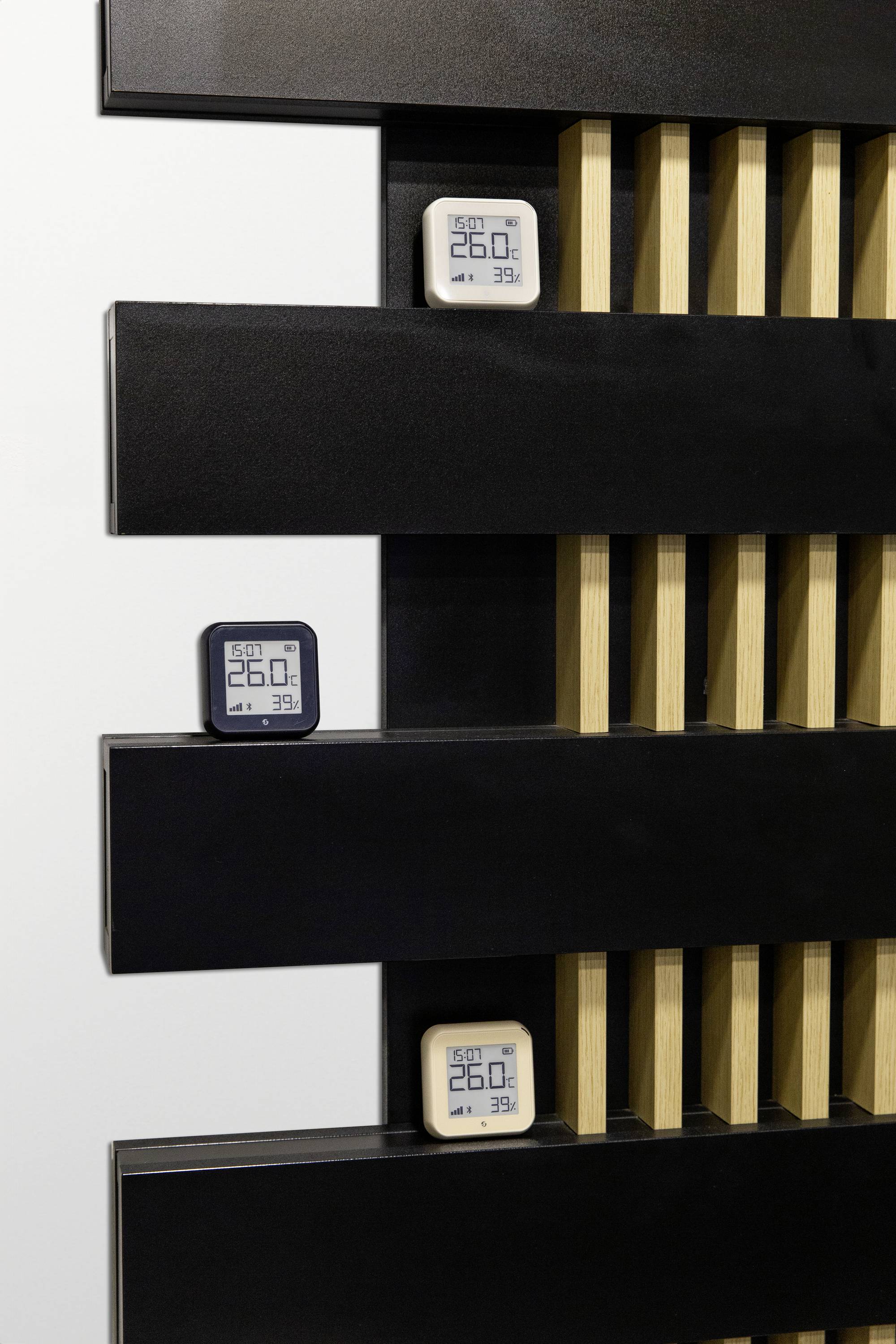Drei digitale Thermo-Hygrometer zeigen 26,0°C und 39% Luftfeuchtigkeit an, montiert an einer Wand mit schwarzen und holzfarbenen Elementen.