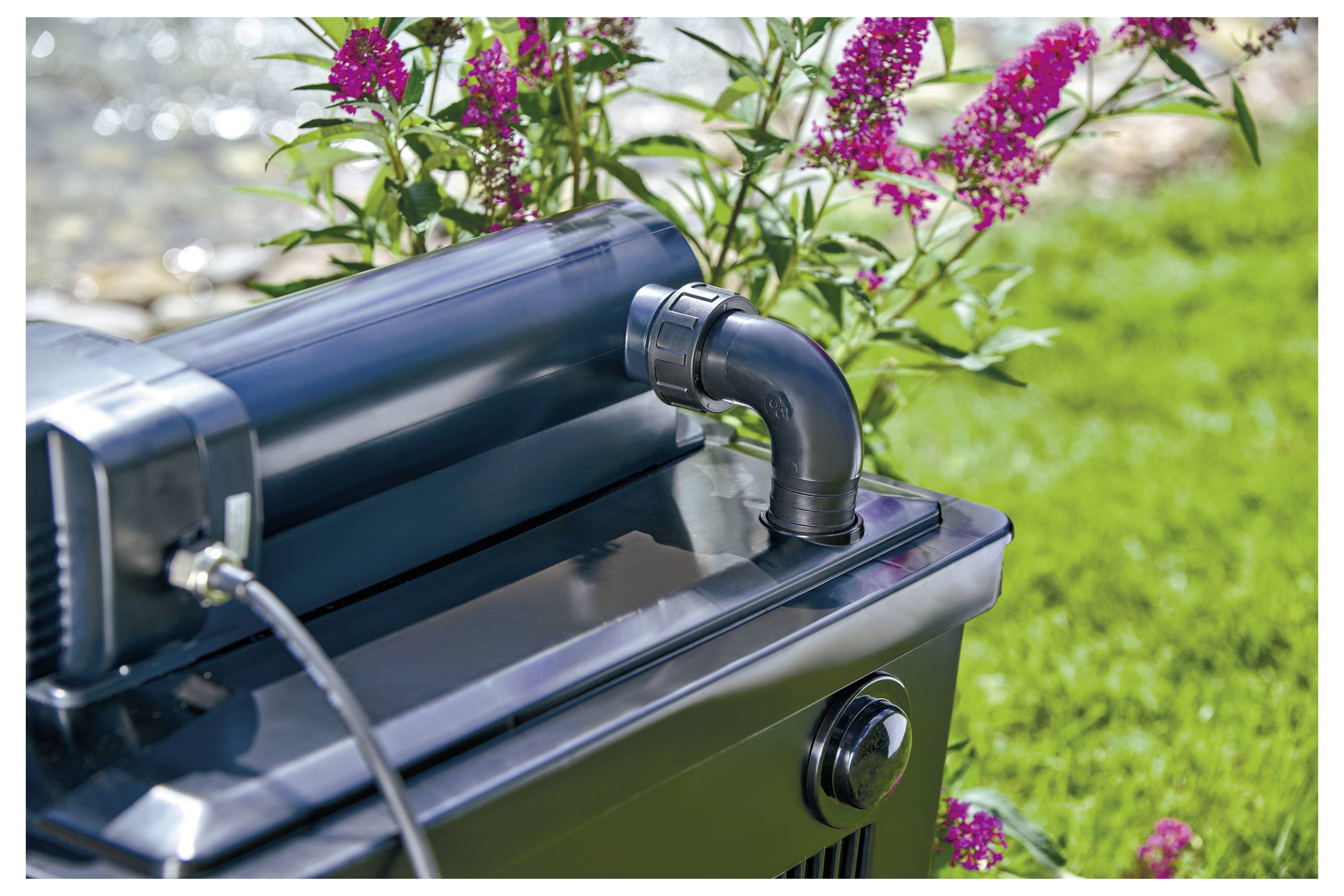Eine schwarze Außenwärmepumpe mit Metallrohranschluss ist in der Nähe von leuchtend pinken Blumen und einem grünen Grashintergrund platziert.