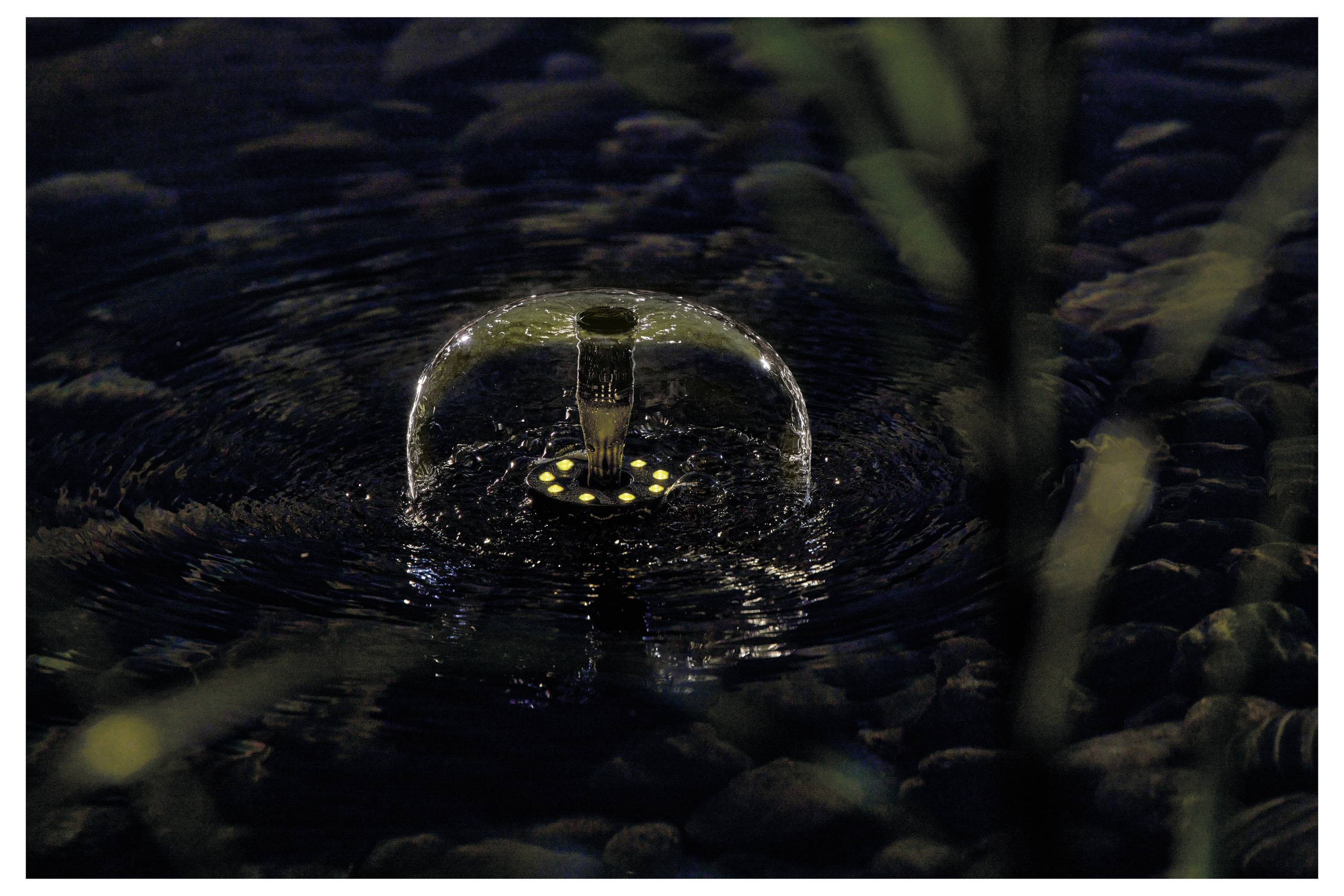 Ein kleiner Springbrunnen-Lichtstrahl erhellt die Nacht und erzeugt Wellen auf einer ruhigen Wasseroberfläche, umgeben von Kieselsteinen und teilweise von Blattwerk verdeckt.