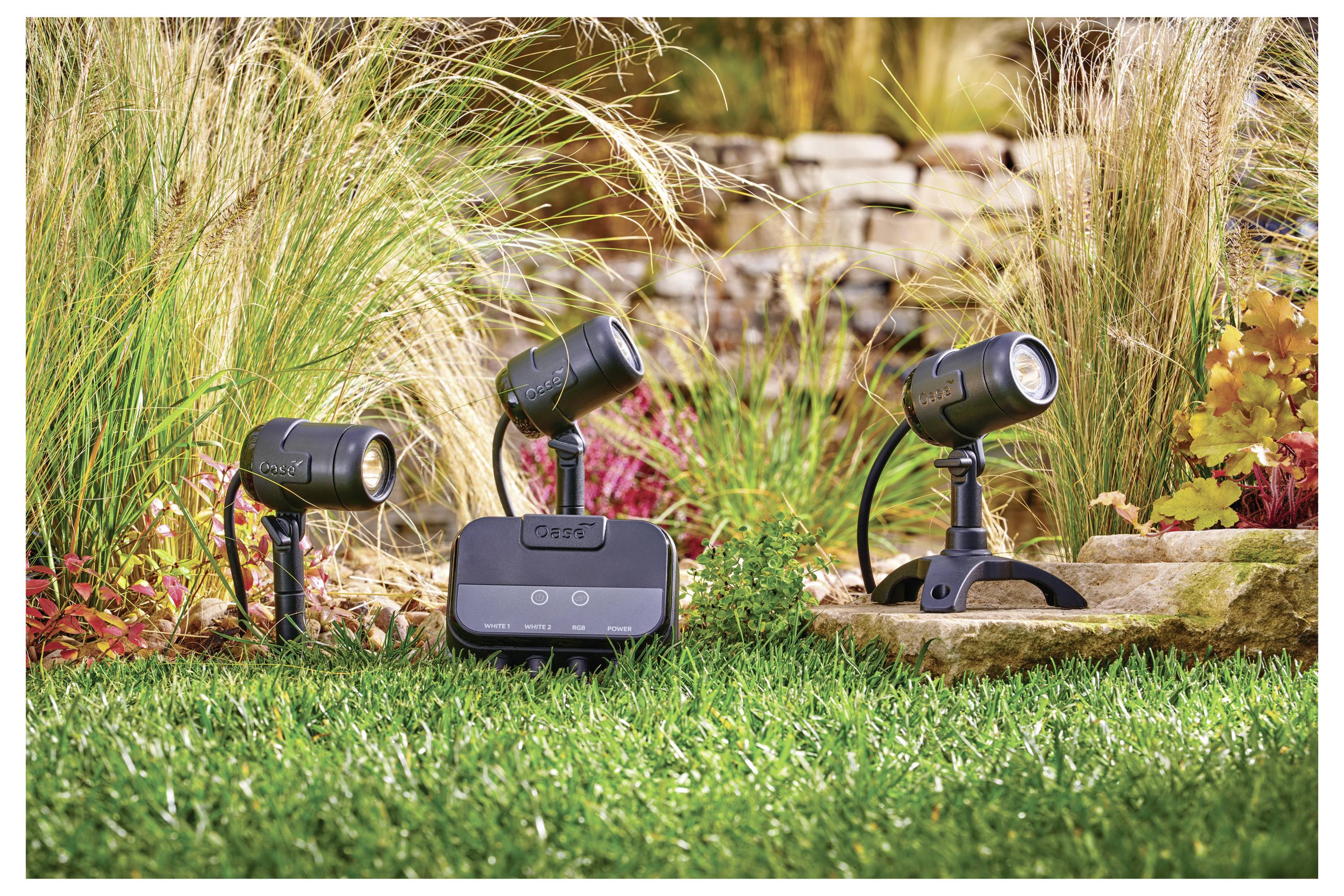 Smarte Außenbeleuchtung und Steuergerät auf Gras, umgeben von Pflanzen und Steinen, präsentiert eine Gartenbeleuchtungsanlage.