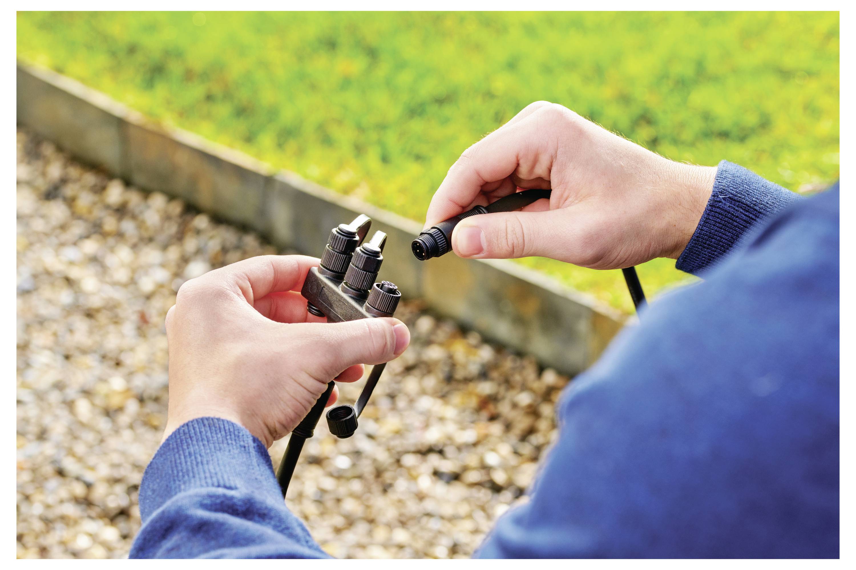 Person in blauem Sweatshirt, der ein Bewässerungssystem in einem felsigen Gartenbereich montiert oder repariert und dabei Verbinder und Rohrleitungen in der Hand hält.