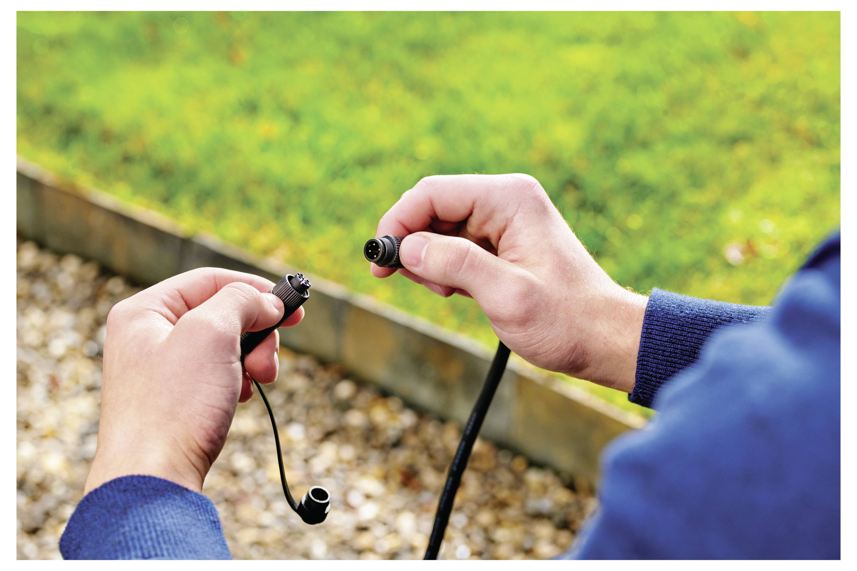 Person, der zwei Gartenschlauchverbindungen zusammenfügt, steht auf einem Kiesweg mit Gras im Hintergrund.