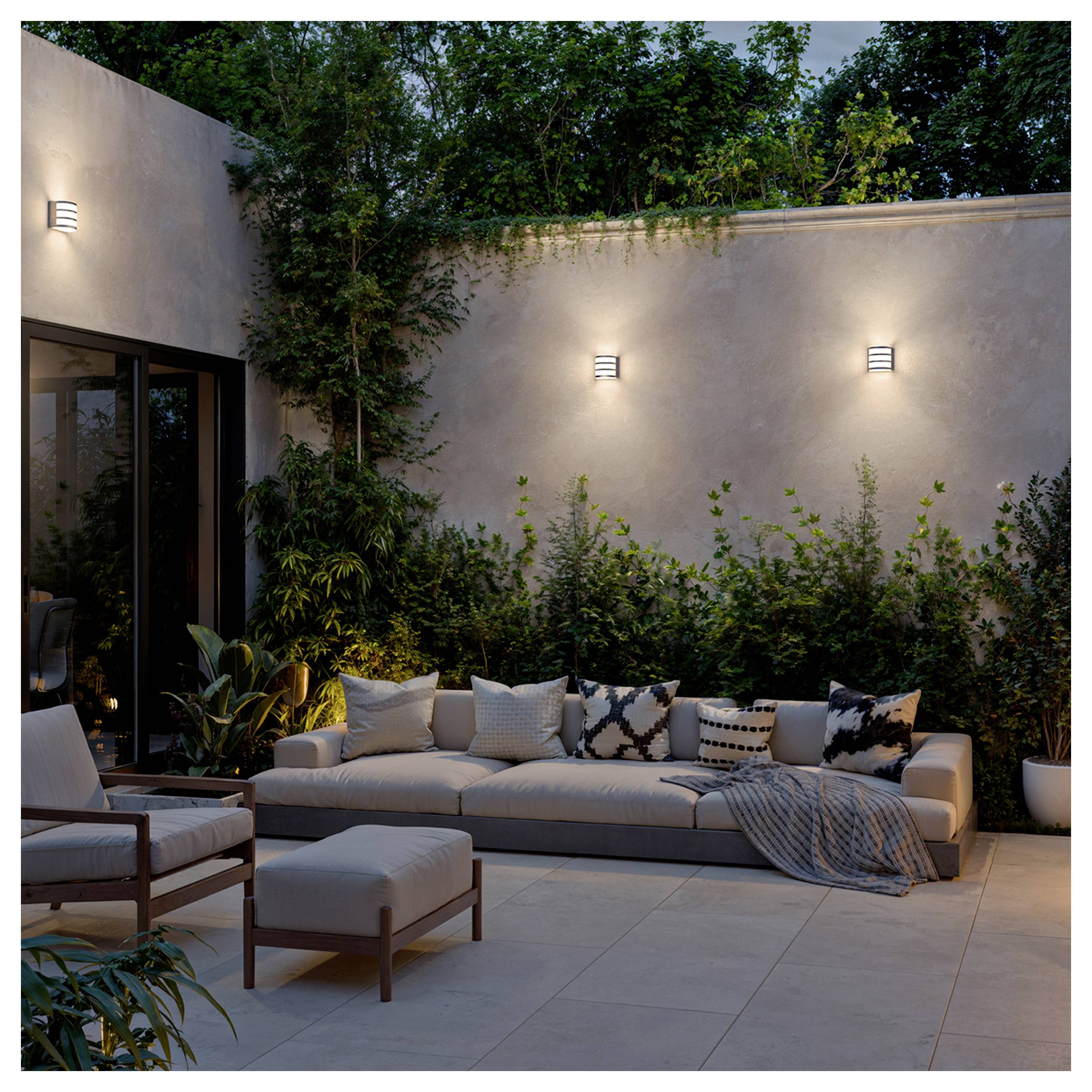 Moderner Außenpatio mit langem Sofa, Loungesessel und Ottomane vor einer beleuchteten Stuckwand, umgeben von Pflanzen in der Dämmerung.