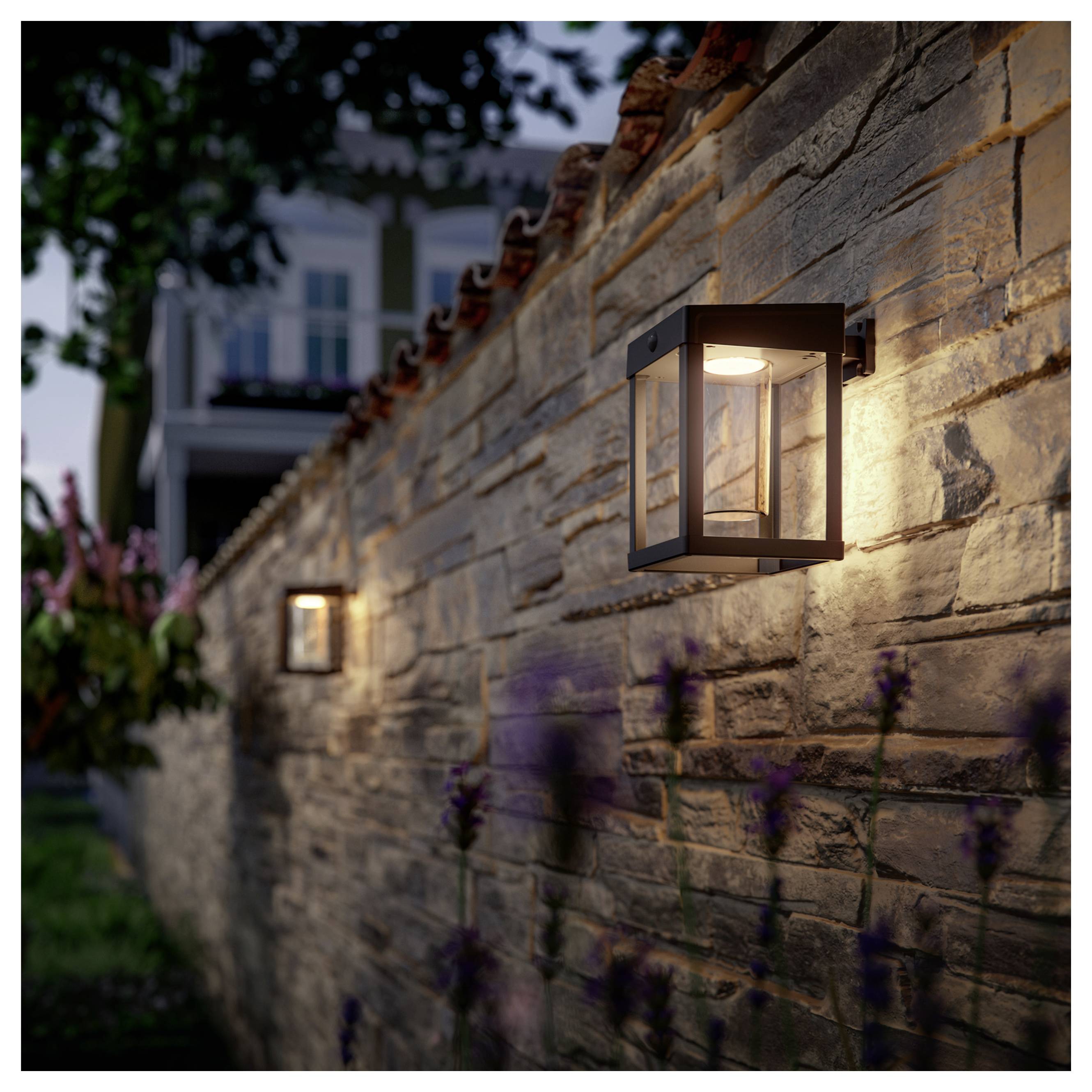 An der Wand montierte Außenlaternen, die in der Dämmerung auf einer steinernen Gartenmauer leuchten, mit einem unscharfen Haus im Hintergrund.