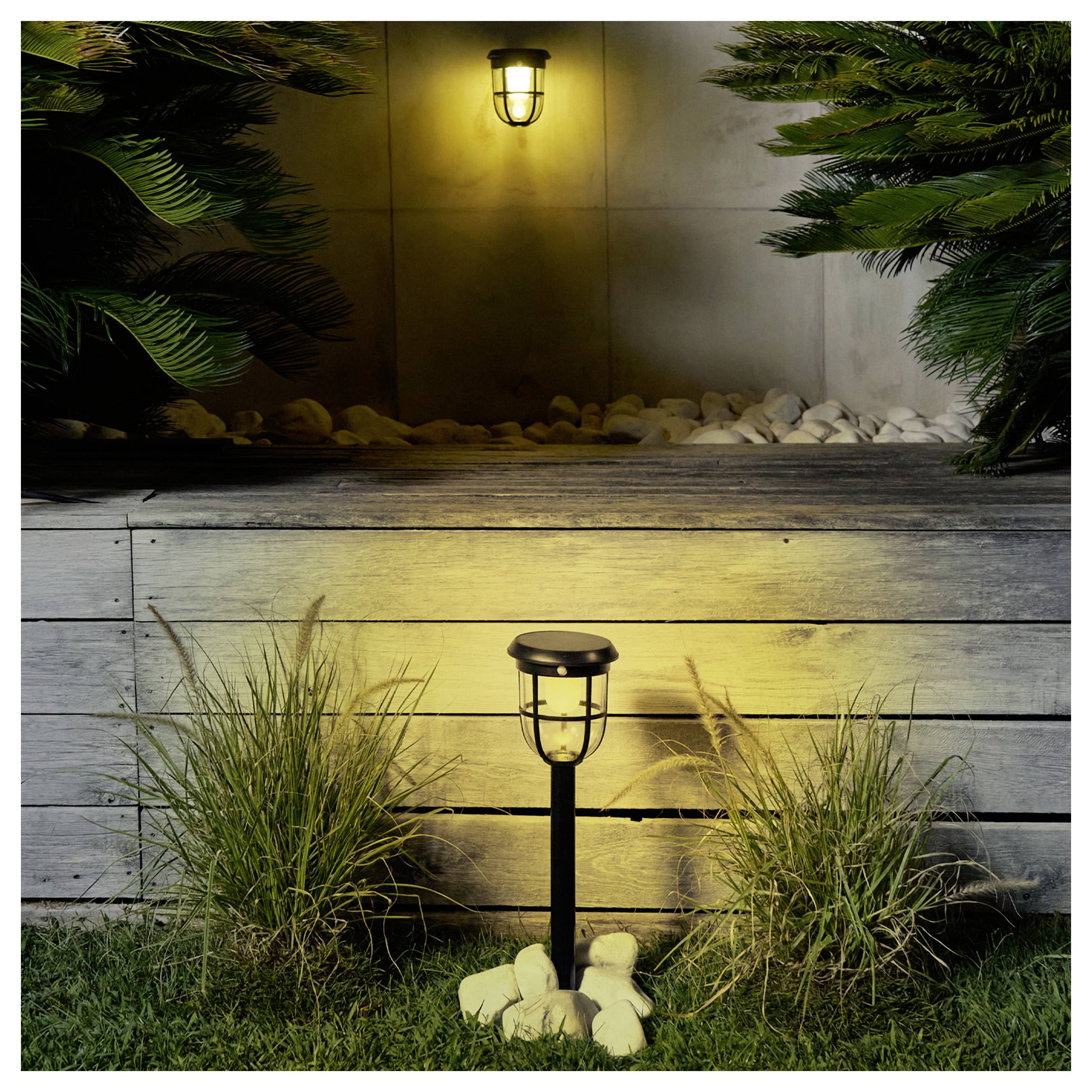 Gartenleuchte im Außenbereich vor einer erhöhten Pflanzerwand mit einer leuchtenden Laterne und dekorativen Pflanzen bei Nacht.
