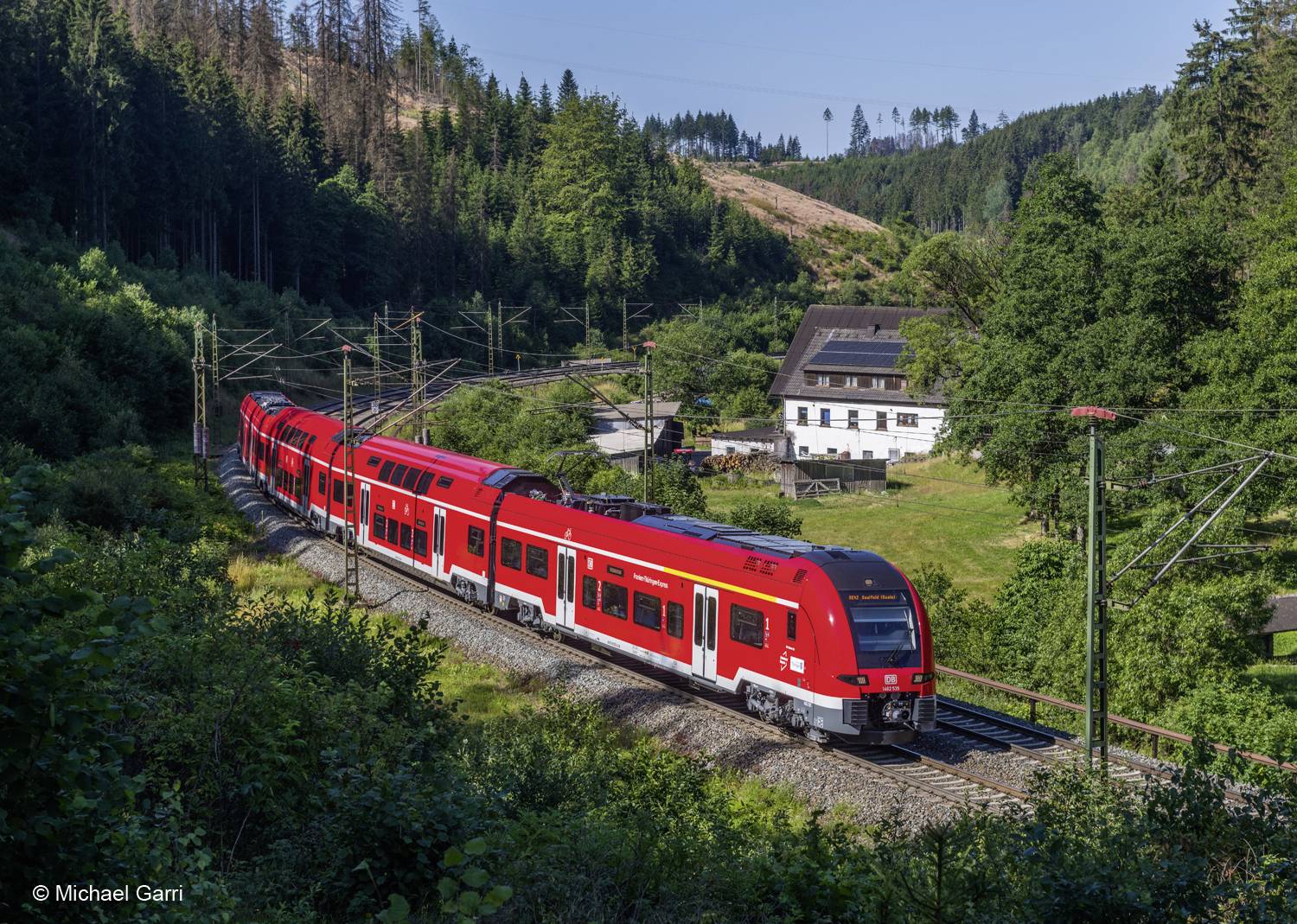 Ein roter Regionalzug fährt tagsüber durch eine waldige Landschaft. Im Hintergrund ist ein weißes Gebäude zu sehen.