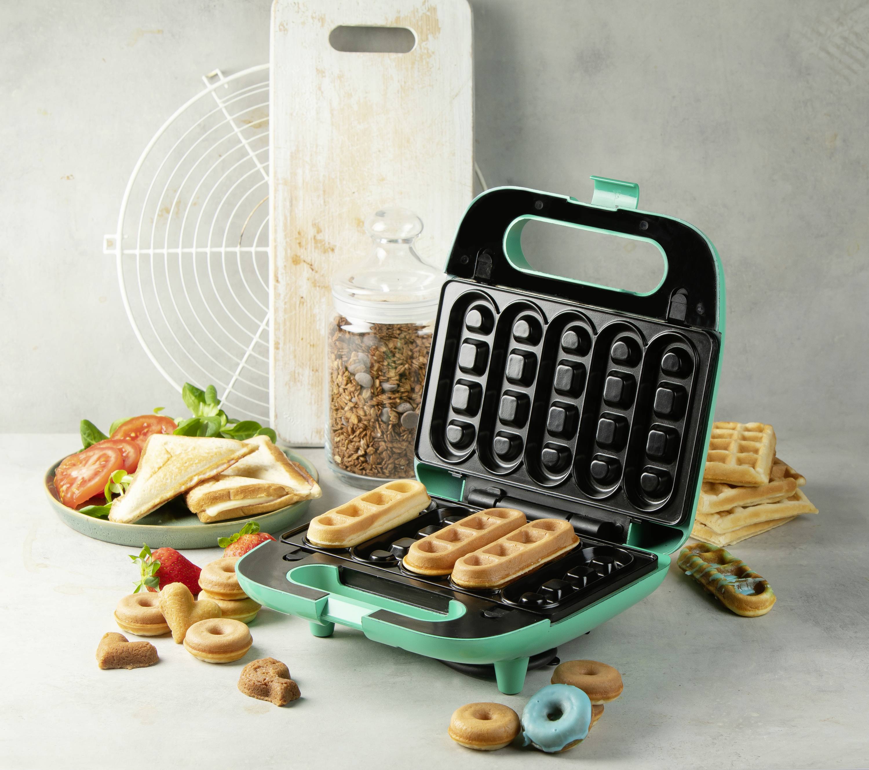 Ein grüner Waffelautomat mit frischen Waffeln und daneben liegendem Gebäck, daneben ein Teller mit Sandwiches und Tomaten.