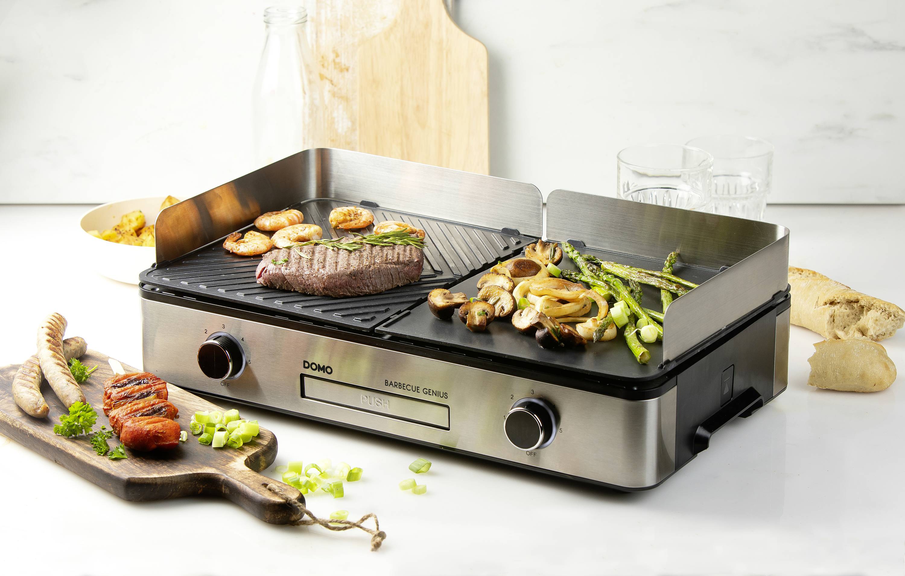 Ein elektrischer Grill mit Fleisch und Gemüse. Steak, Spargel, Pilze und Zwiebeln liegen auf dem Grill. Daneben Brot und Kräuter.