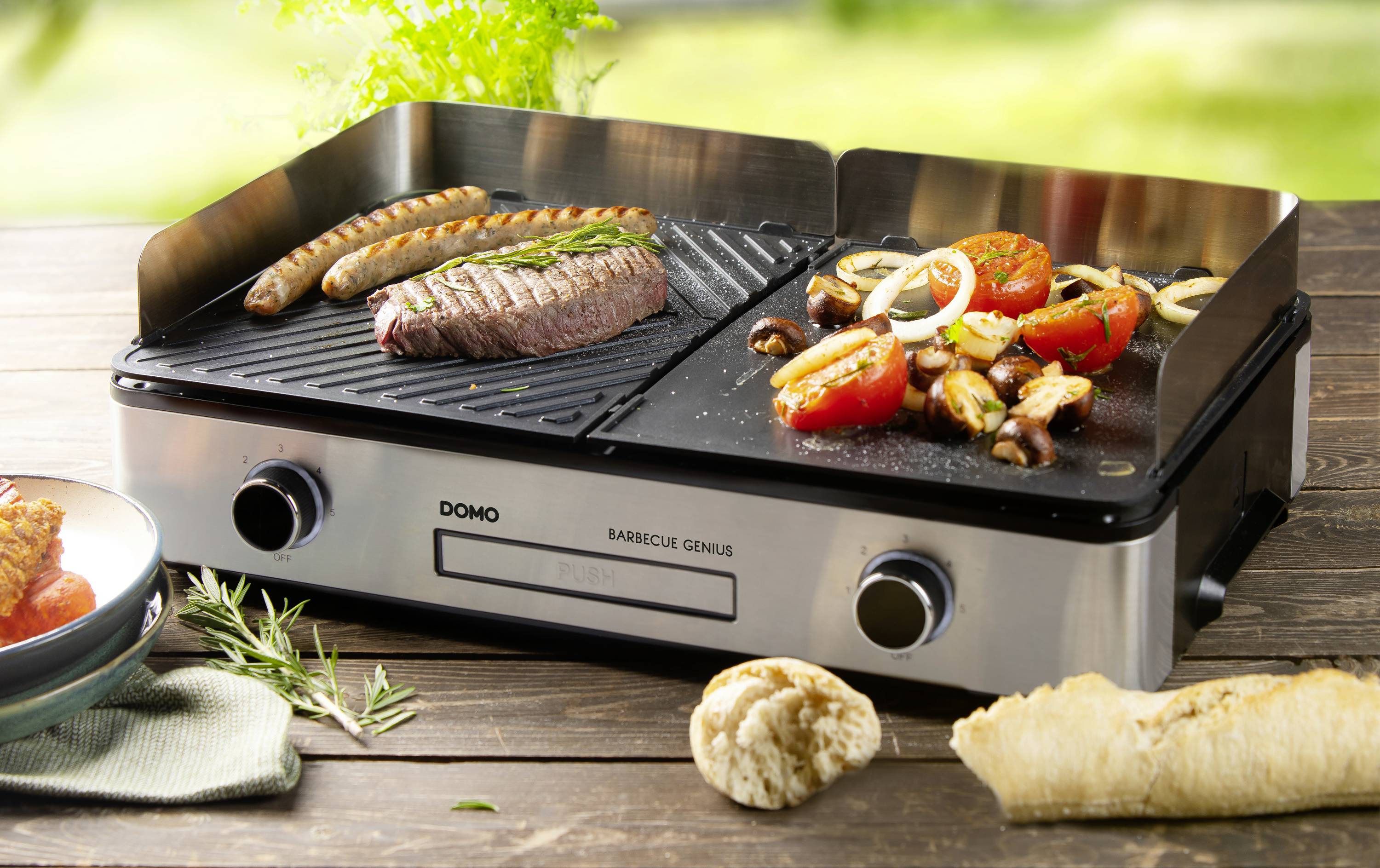 Elektrogrill mit Bratwurst, Steak und Grillgemüse wie Tomaten und Pilze auf zwei Grillplatten. Frisches Brot und Kräuter daneben.