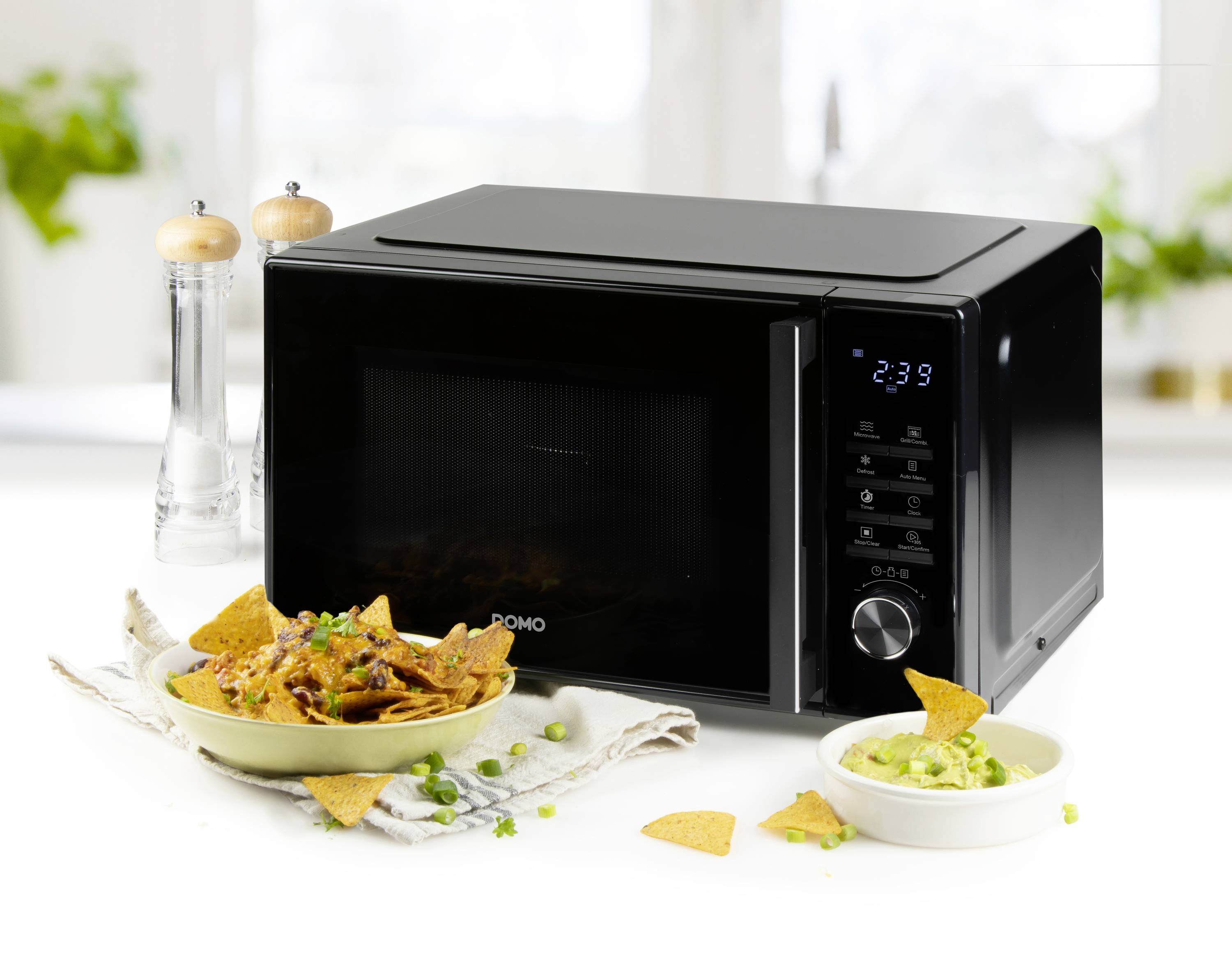 Schwarze Mikrowelle auf weißem Tisch, umgeben von Pfefferstreuer, Salzstreuer und Teller mit Nachos und Guacamole.