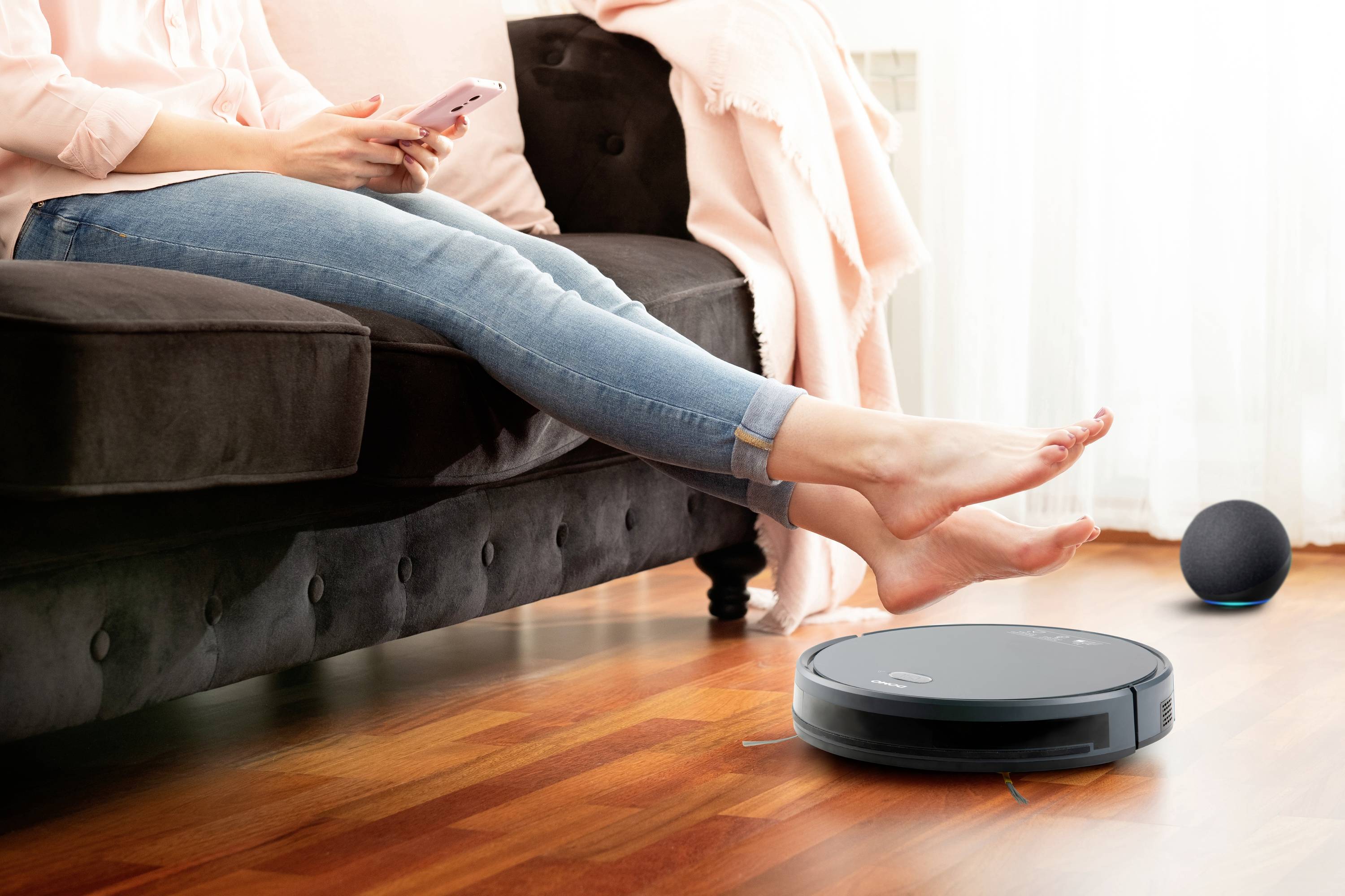 Eine Person sitzt auf einem Sofa mit ausgestreckten Beinen und hält ein Smartphone. Ein Roboterstaubsauger reinigt den Holzboden.