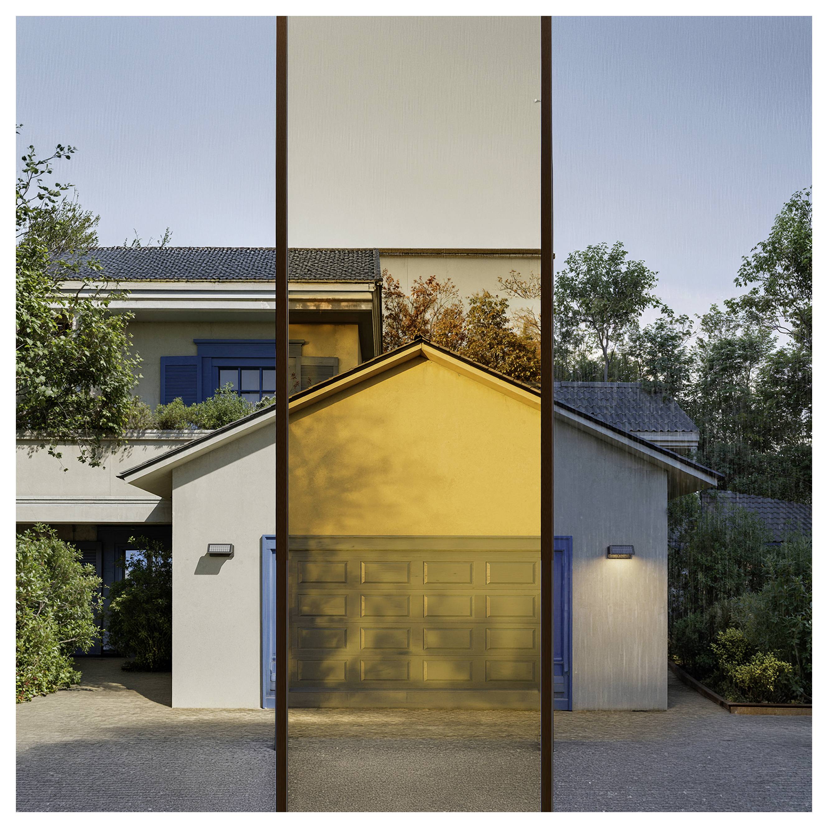 Modernes Haus, das durch ein hohes, schmales reflektierendes Panel zu sehen ist, mit Sonnenlicht, das in der Abenddämmerung auf die Garage und die umgebenden Bäume reflektiert wird.