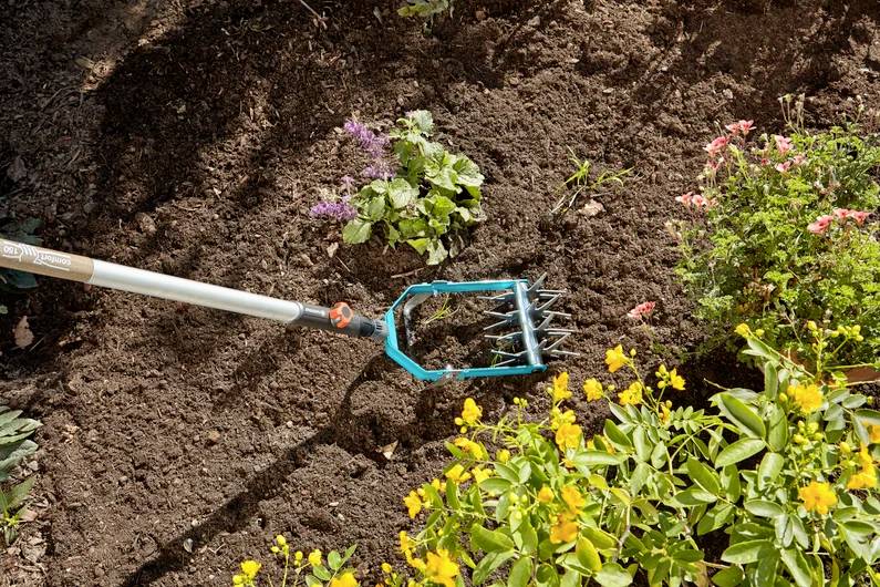 Ein Gartengerät lockert den Boden in einem Blumenbeet mit leuchtend gelben, rosa und lila Blumen und verbessert so die Gartenkultivierung.