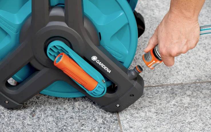 Eine Person befestigt einen Gartenschlauch an einer Gardena-Schlauchtrommel auf einer gefliesten Oberfläche und bereitet sich auf Bewässerungsaufgaben im Freien vor.