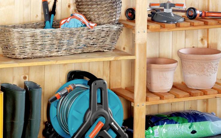 Gartenhaus mit Gartengeräten, aufgerolltem Schlauch und leeren Blumentöpfen auf Holzregalen. Gummistiefel stehen am Boden.