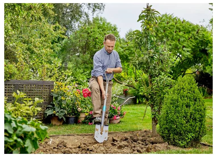 Eine Person in einem Garten pflanzt einen Baum mit einer Schaufel. Die Umgebung ist üppig mit verschiedenen Pflanzen und Blumen, was einen Fokus auf Gartenbau andeutet.