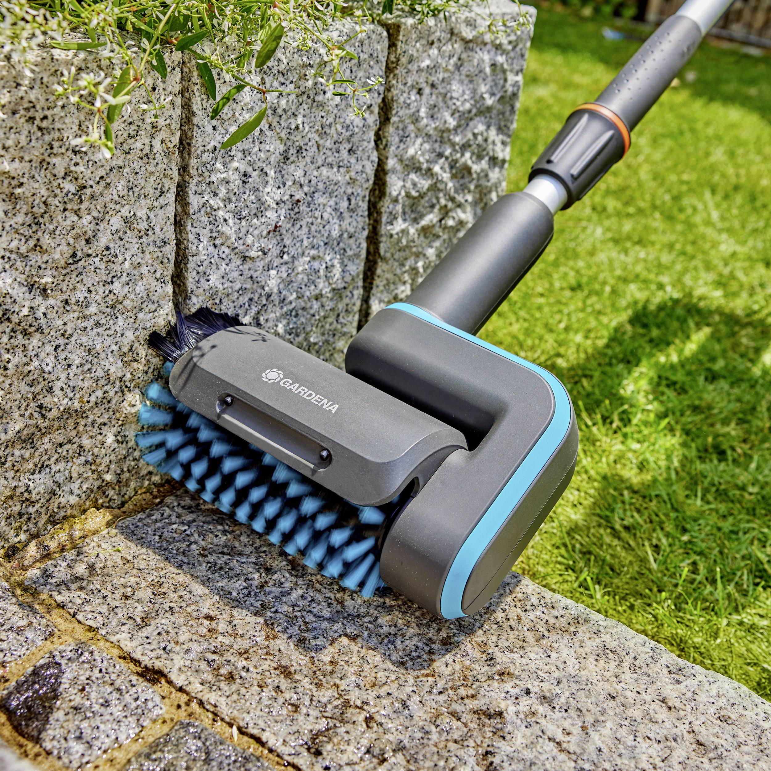 Bürstenreinigungsgerät mit blau-grauen Borsten reinigt die Kante eines gepflasterten Wegs neben einem grünen Rasen.