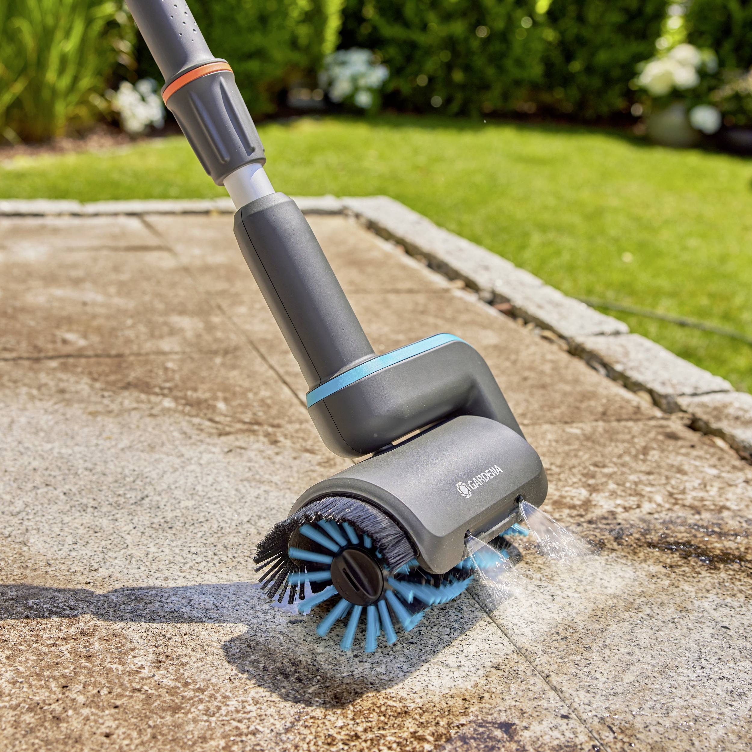 Ein elektrisch betriebener Terrassenreiniger reinigt einen mit Steinplatten belegten Weg im Garten. Im Hintergrund sind grüne Büsche.