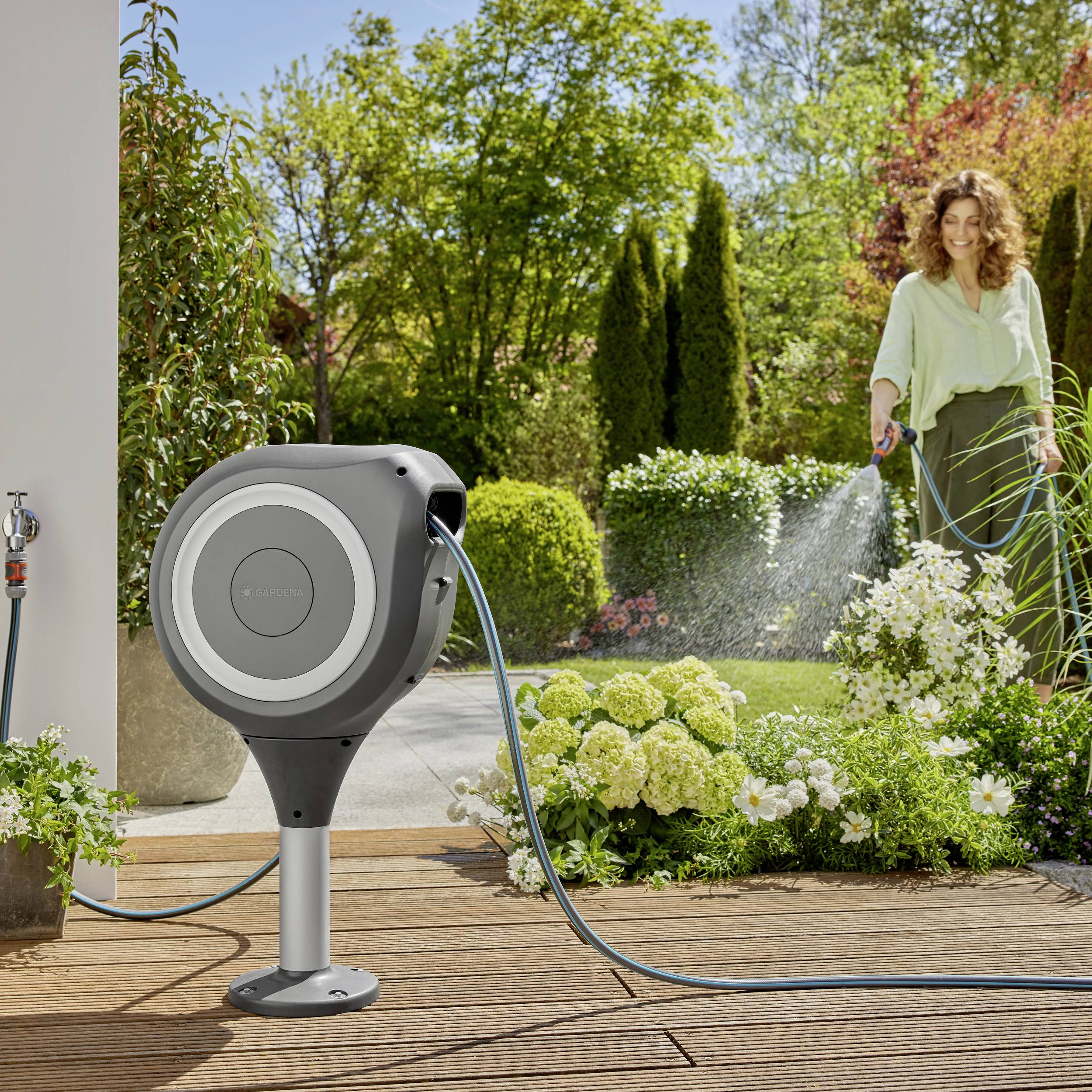 Eine Frau gießt Pflanzen in einem Garten mit einem Wasserschlauch aus einer modernen, runden Gartenspritze. Helder Tag, viel Grün.