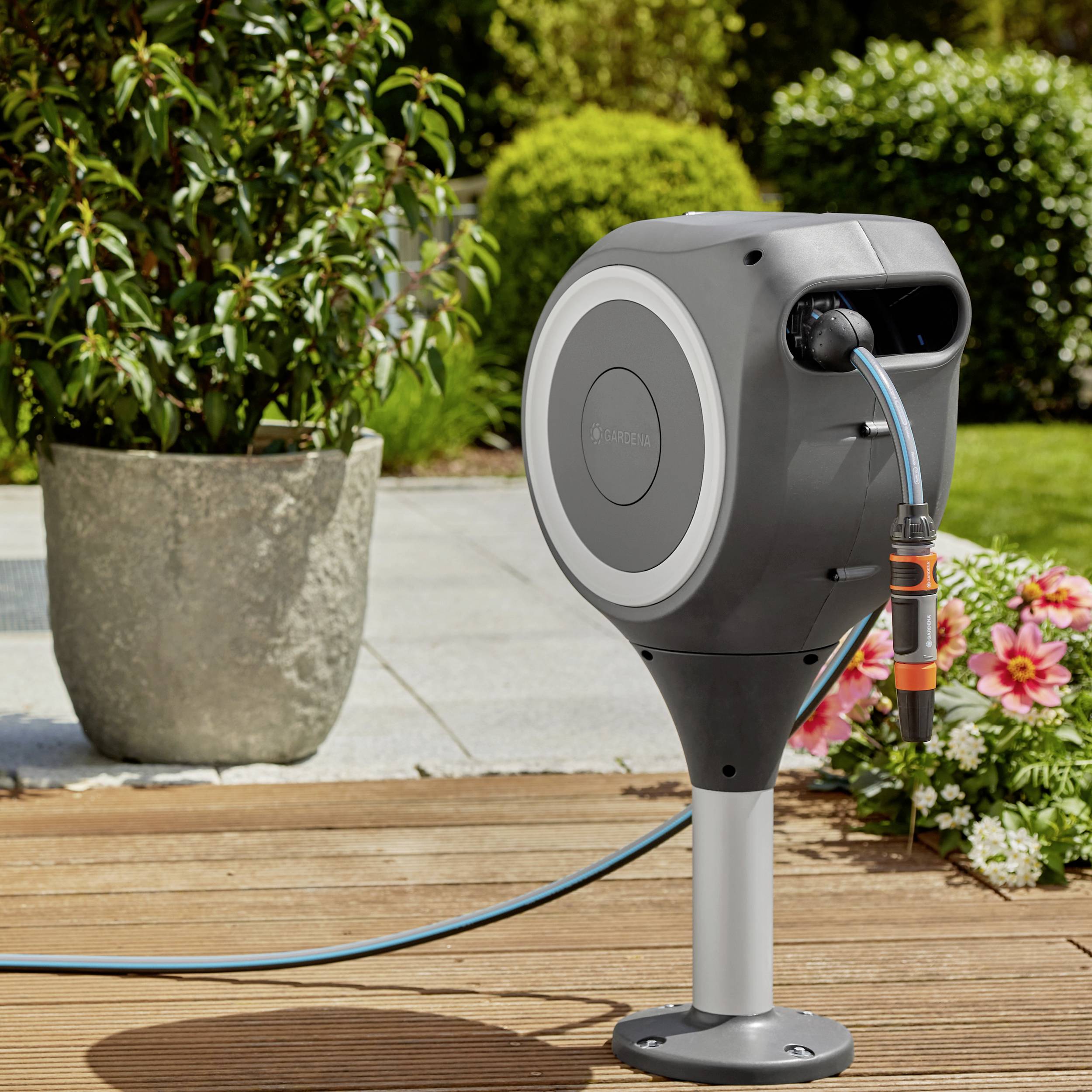 Eine graue Gartenschlauchtrommel mit automatischem Aufrollmechanismus steht auf einer Terrasse neben Blumen und einem großen Pflanzgefäß.