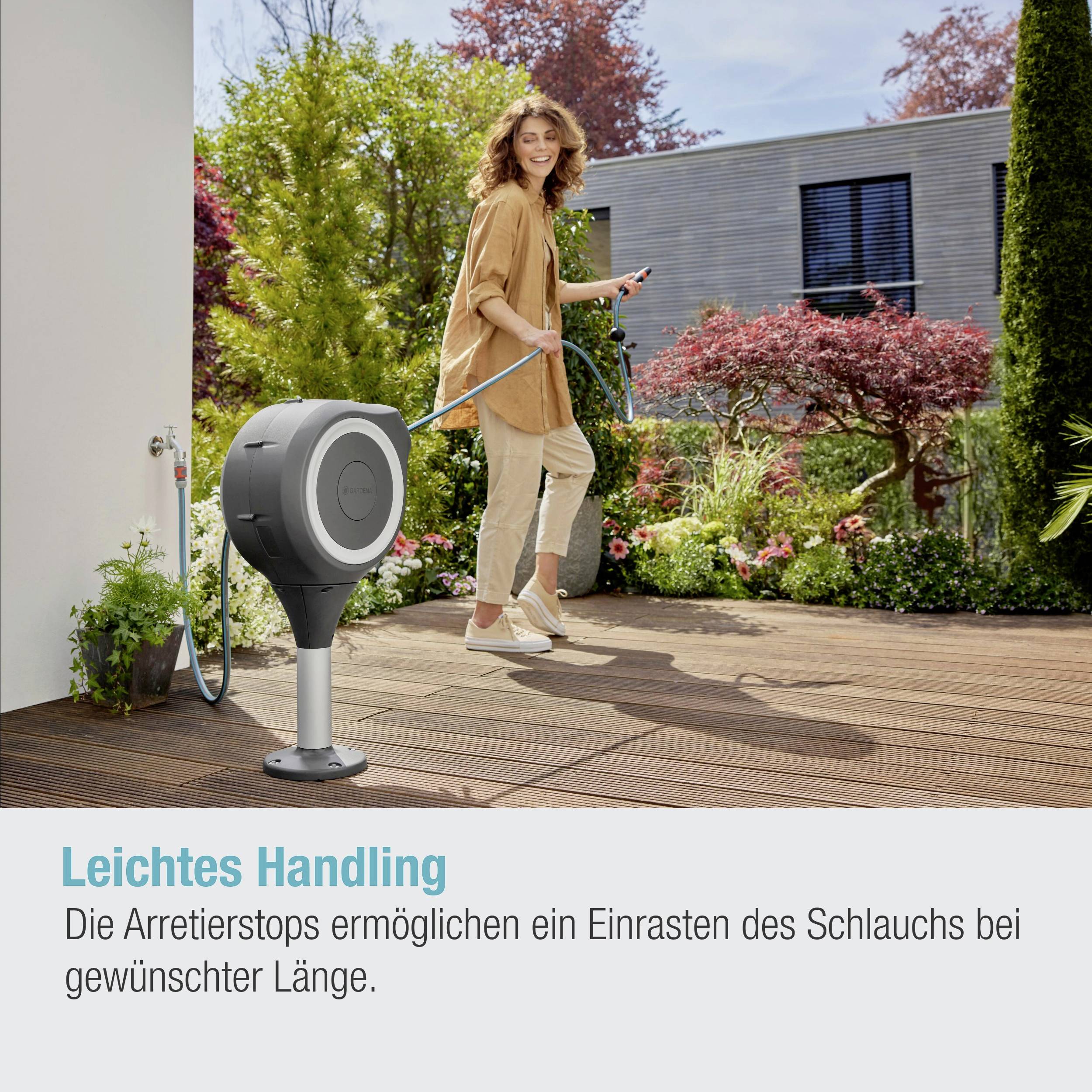 Eine Person steht im Garten, hält einen ausziehbaren Wasserschlauch, der an einer grauen Wandhalterung befestigt ist. Bunte Pflanzen im Hintergrund.