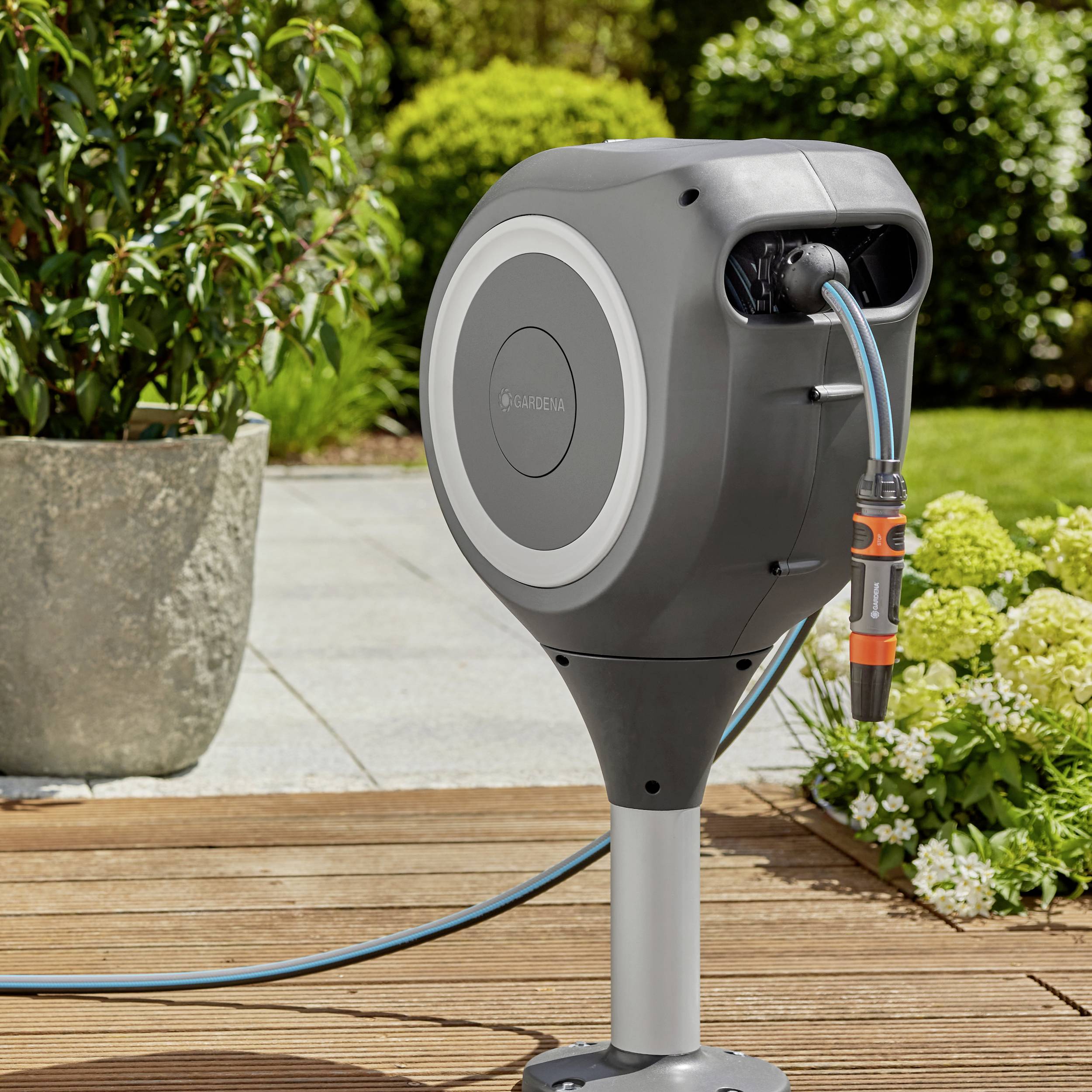 Ein automatischer Gartenschlauch auf einer Halterung steht im Garten. Im Hintergrund sind Pflanzen und Pflastersteine zu sehen.