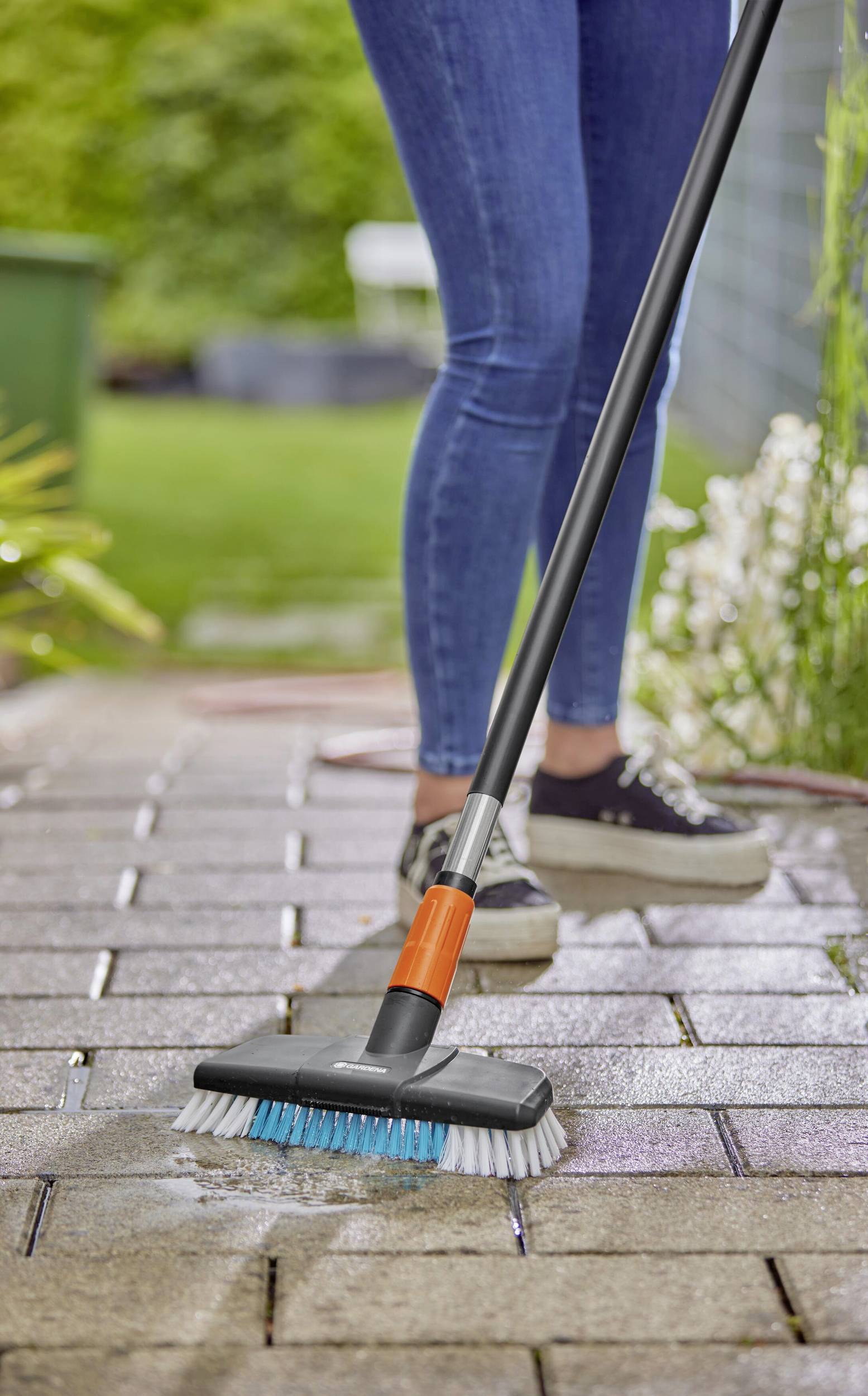 Eine Person reinigt mit einem Schrubber eine gepflasterte Terrasse. Im Hintergrund sind Pflanzen und ein Gartenbereich sichtbar.