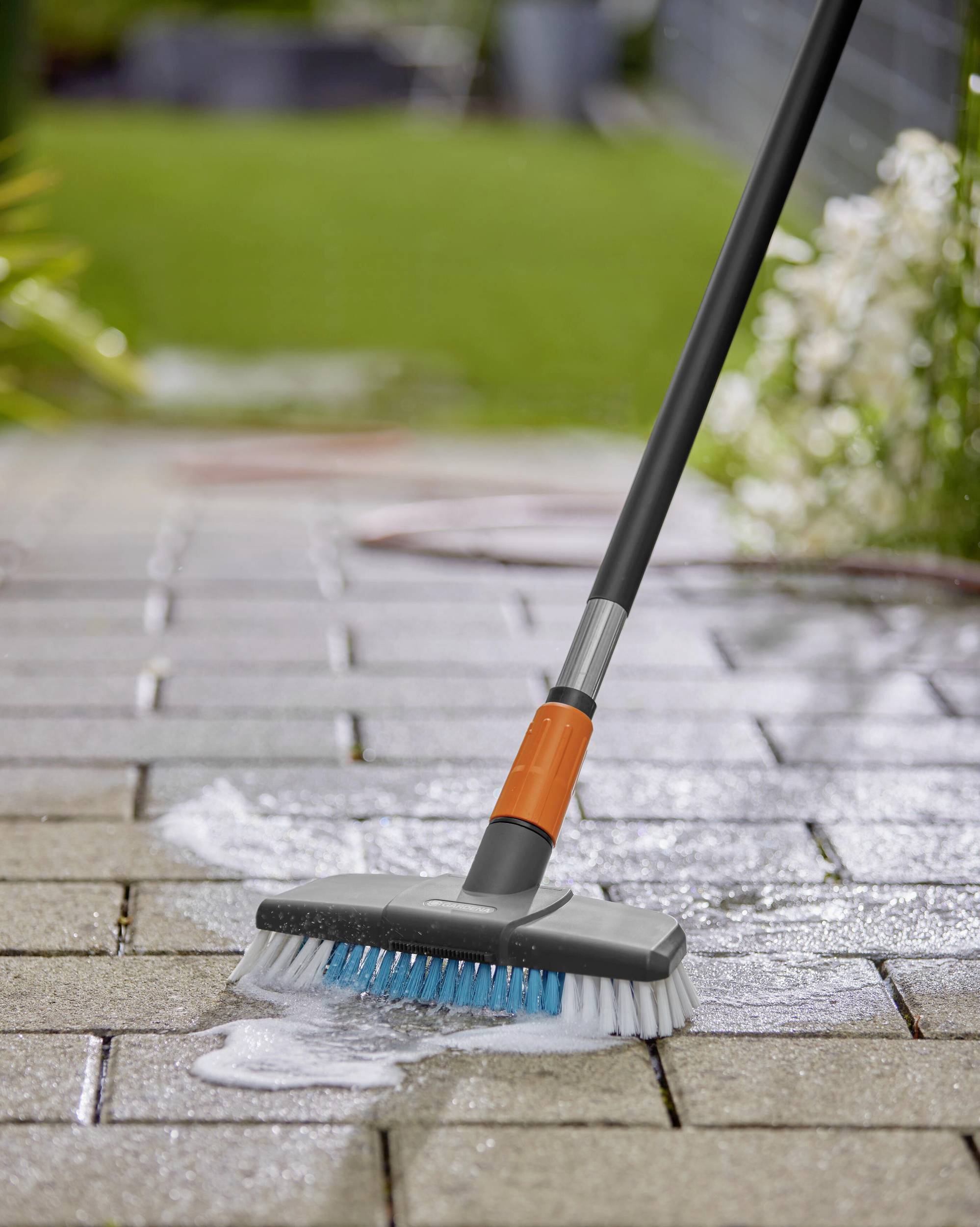 Ein Reinigungsbesen mit orangefarbenem Griff schrubbt eine gepflasterte Terrasse. Seifenwasser bildet schäumende Spuren auf den Fliesen.
