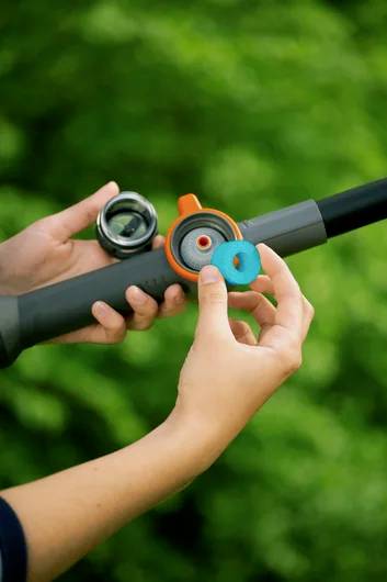 Zwei Hände halten einen Gartenschlauch mit einer Sprühdüse und justieren eine orange-blaue Anschlussvorrichtung vor einem unscharfen, grünen Blätterhindergrund.