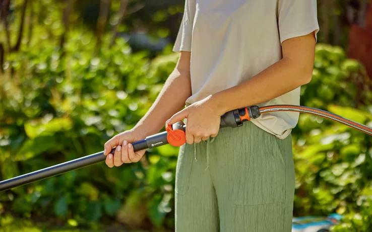 Eine Person hält einen Wasserschlauch in einem Garten und bereitet sich darauf vor, Pflanzen zu gießen. Der Hintergrund ist üppig grün und lässt einen sonnigen Tag vermuten.