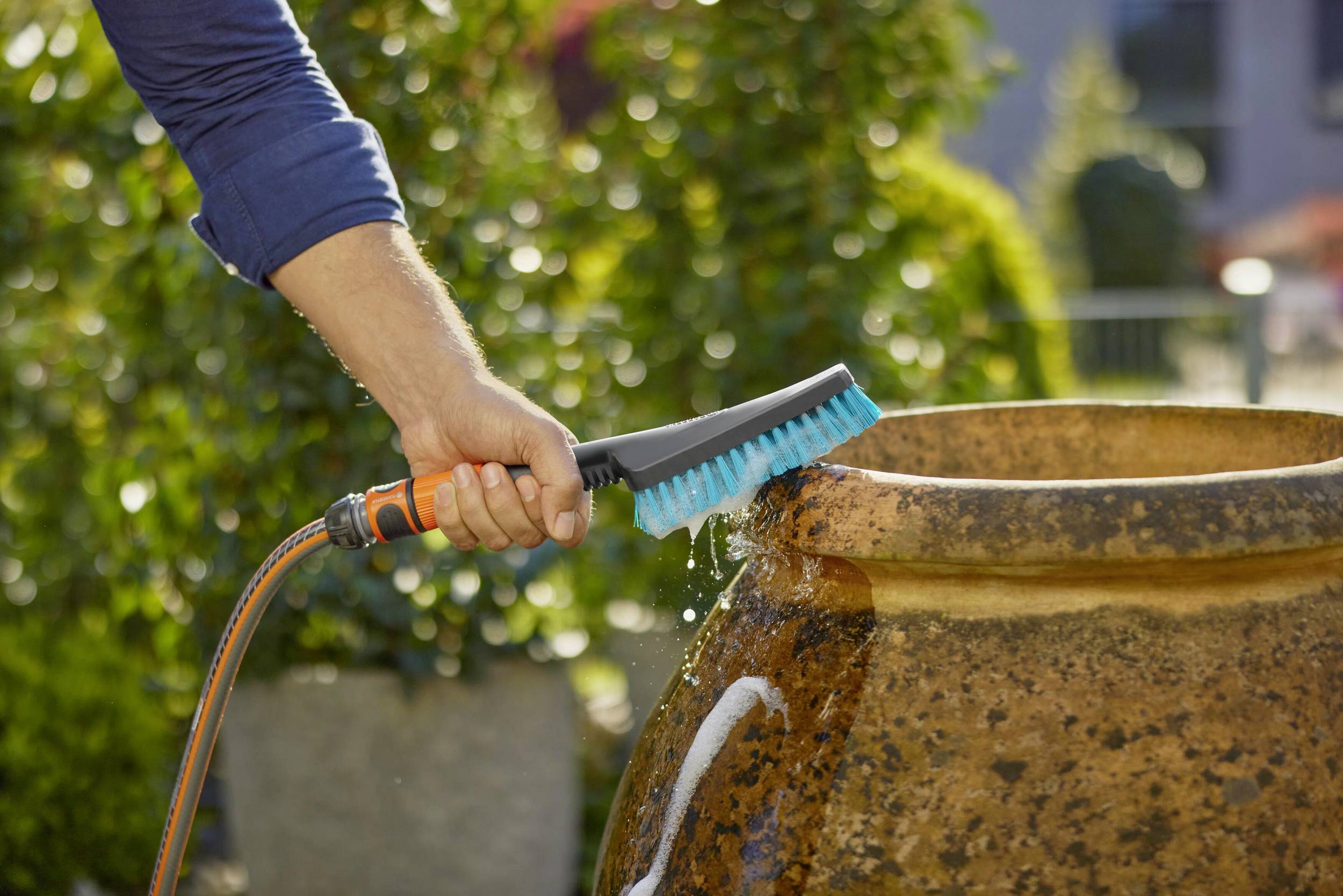 Eine Person reinigt einen großen, runden Keramiktopf im Freien mit einer Wasserbürste. Hintergrund unscharf mit Büschen.